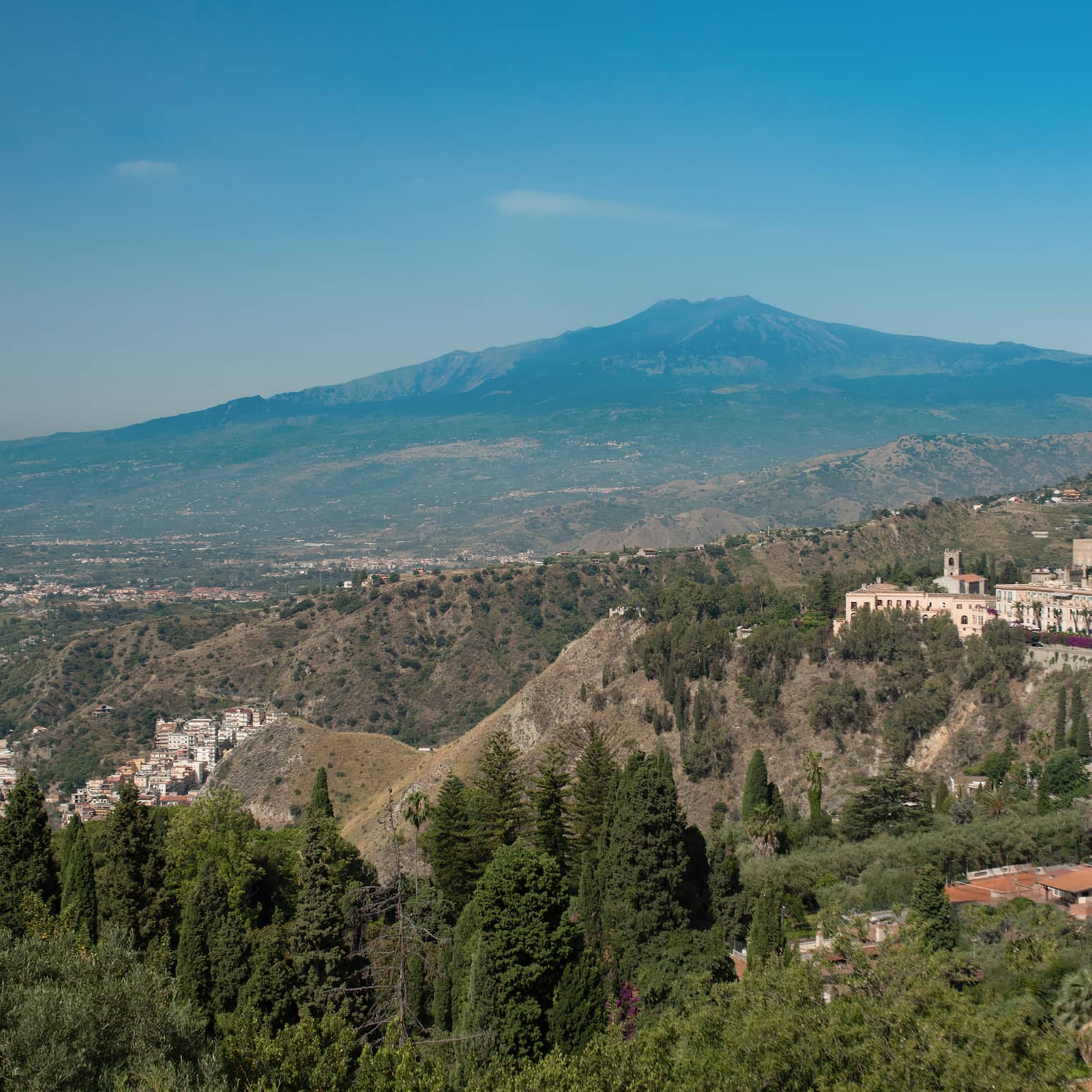 Panoramic view of a coastal town nestled in the hills, overlooking the sea, with mountains in the background and greenery in the foreground.