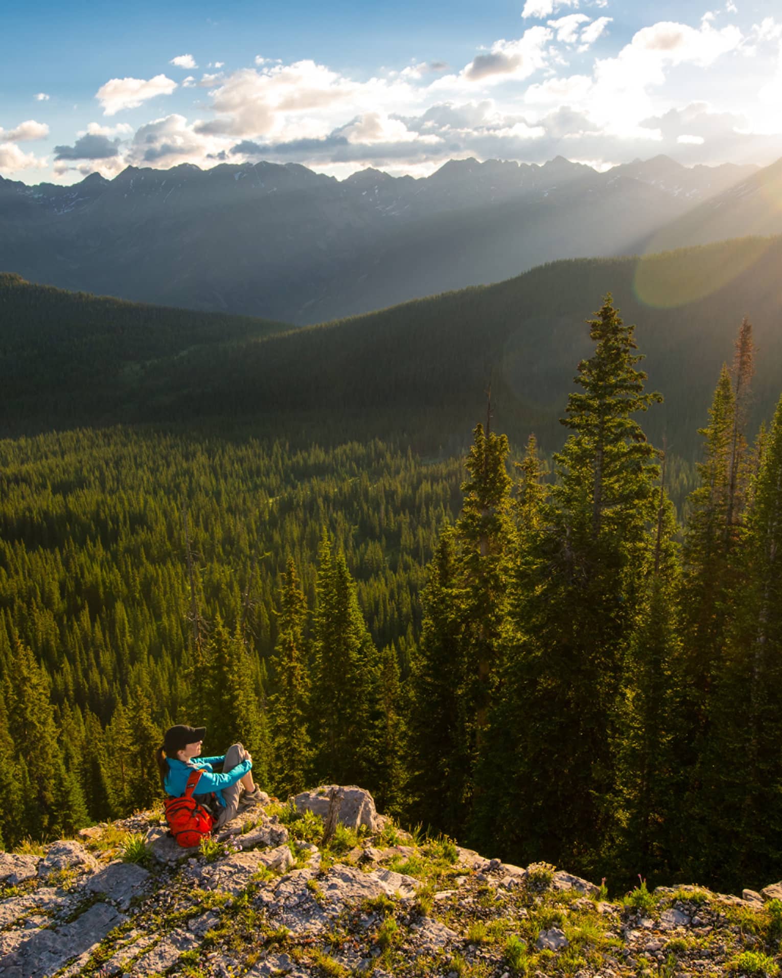 Person sits at edge of cliff overlooking trees, mountains as sun starts to set