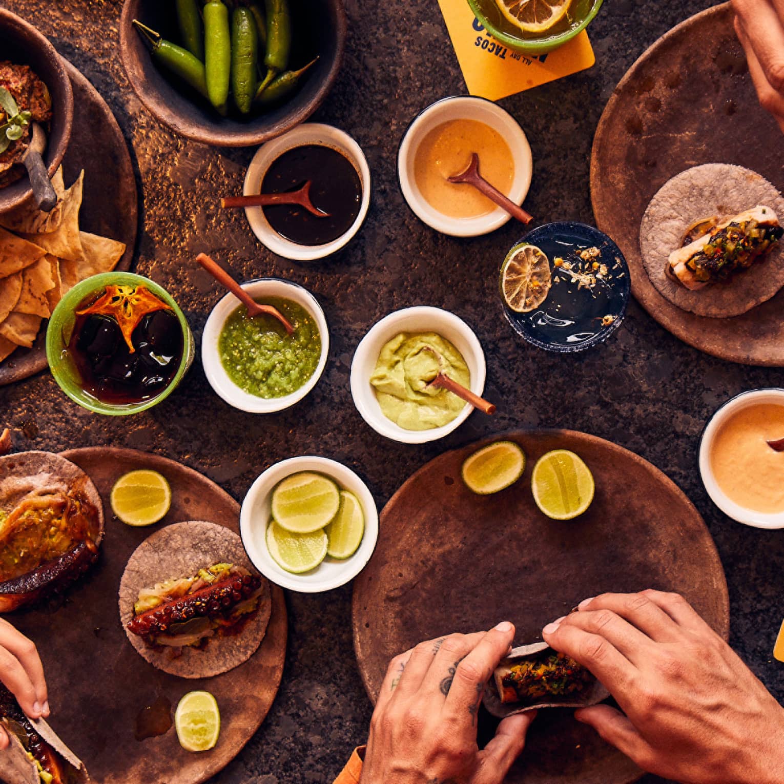 An overhead view of five pairs of hands holding tacos next to bowls of chips, salsa, peppers, limes and sauces.