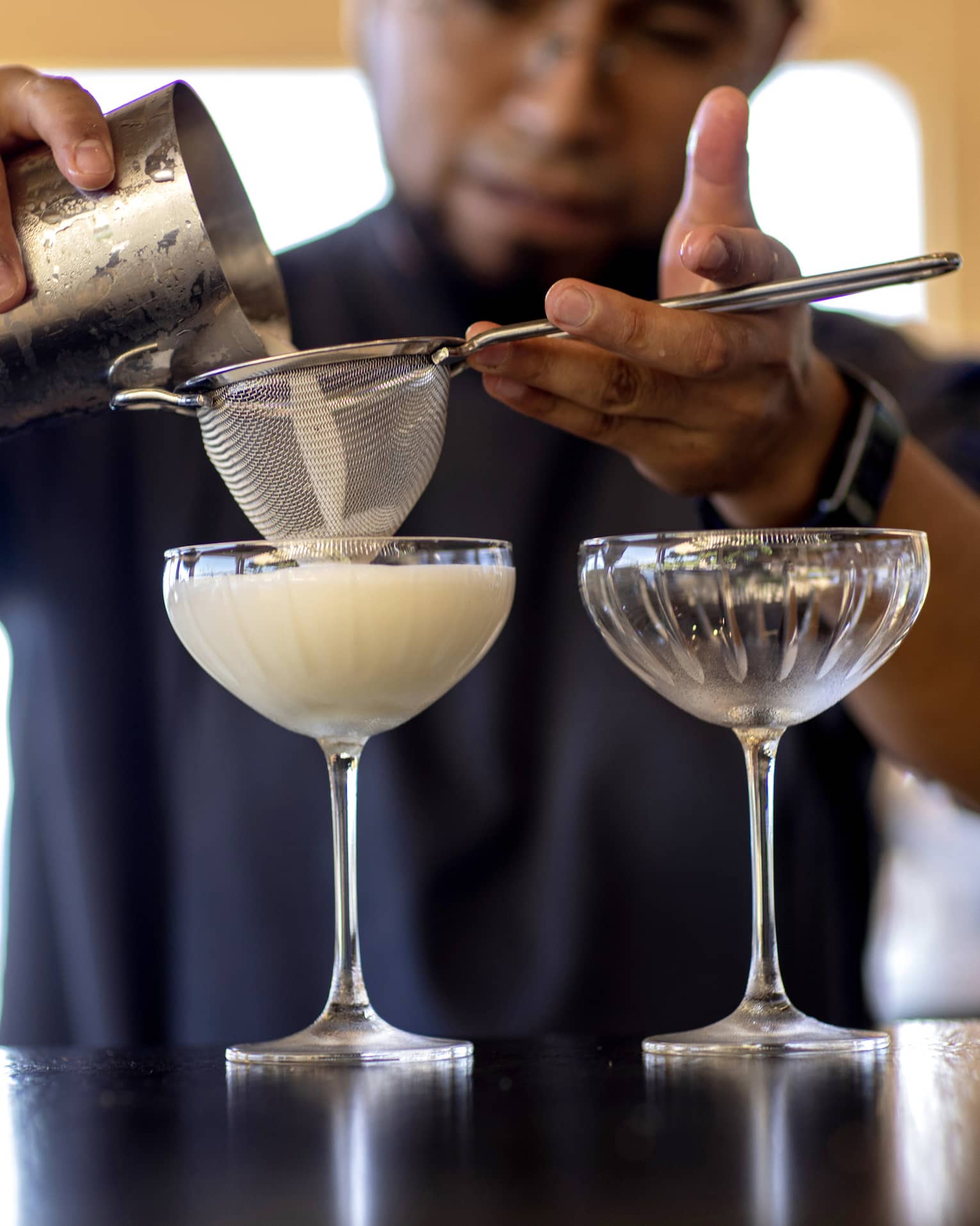 From a metal cocktail shaker, a bartender filters a milky cocktail through a sieve into an etched groove coupe glass. 