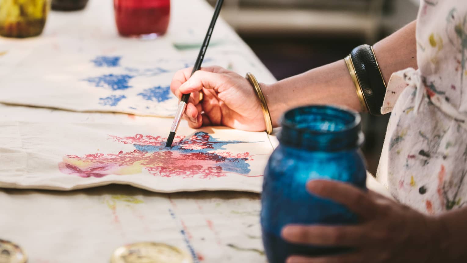 An artist using mason jars filled with red, blue, yellow and black paint to paint images of flowers on canvas tote bags.