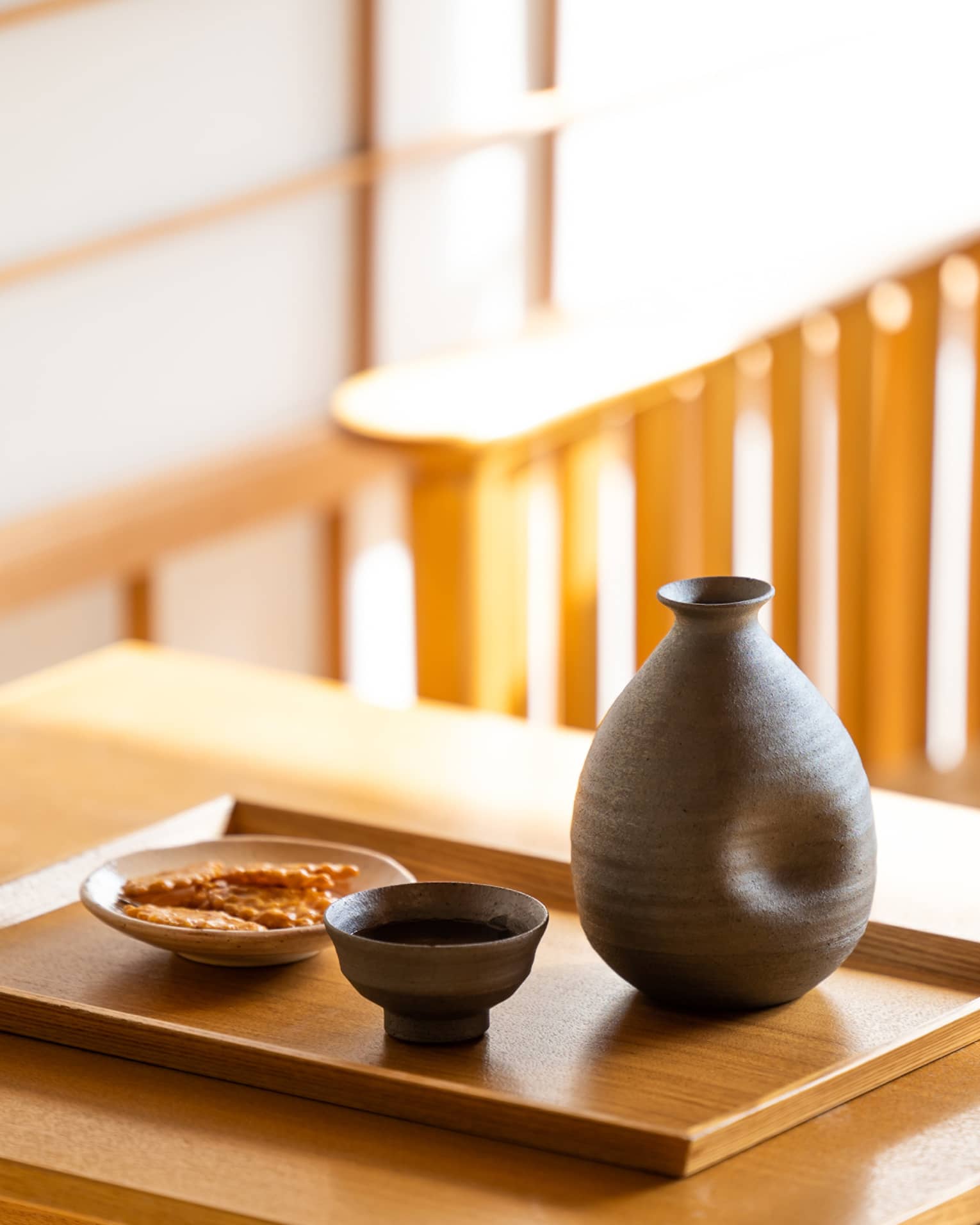 Close-up of a dark ceramic sake carafe with matching cup and a small plate of crackers on a wooden tray and table.