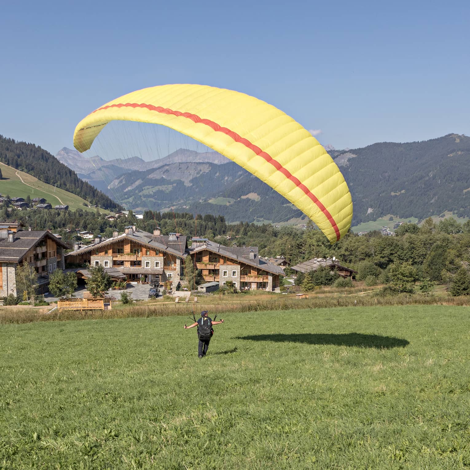Person on meadow holding yellow parachute in air, facing rustic Megève hotel and verdant mountains