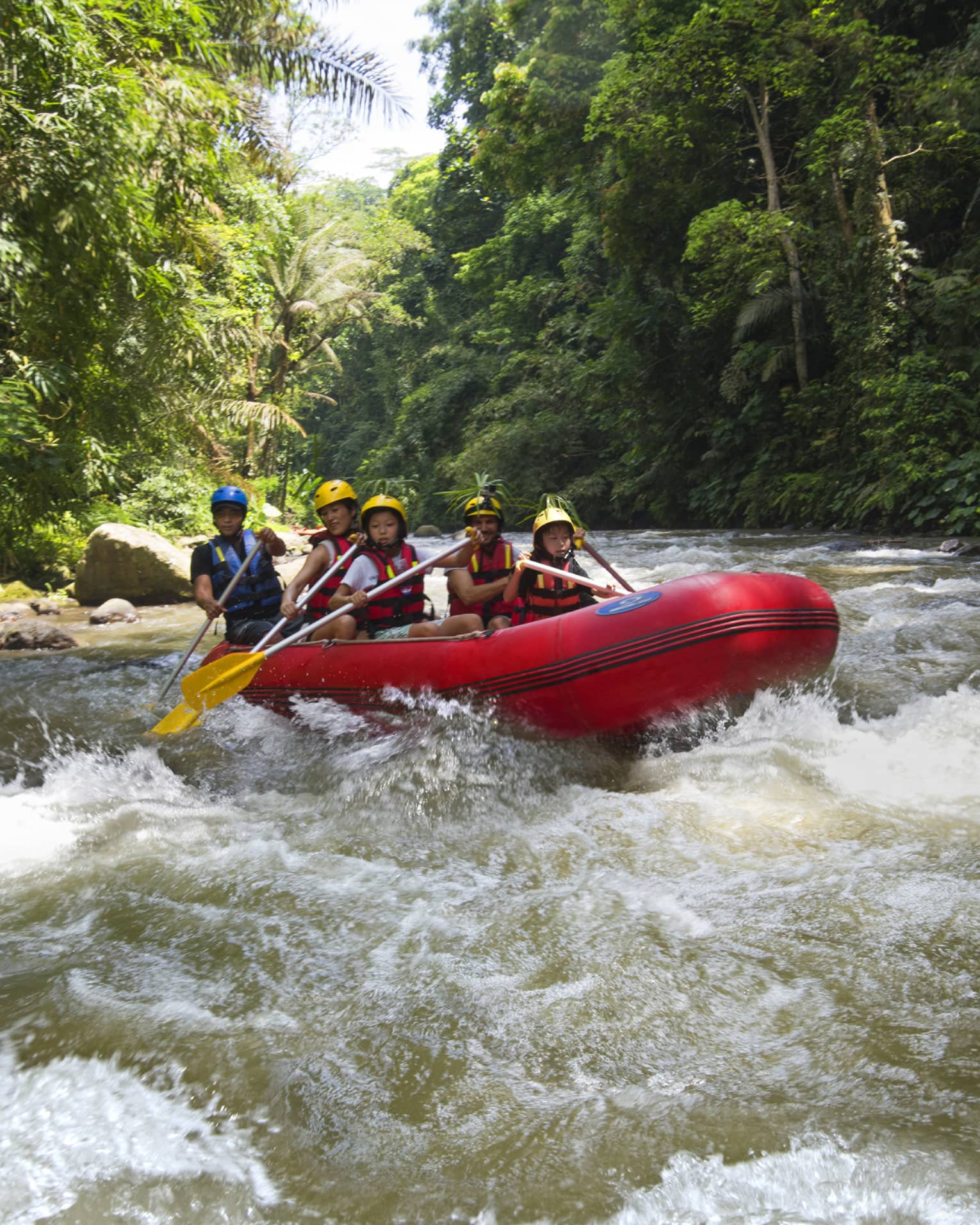 Family of four and guide hold paddles, steer red inflatable raft on rapids in river