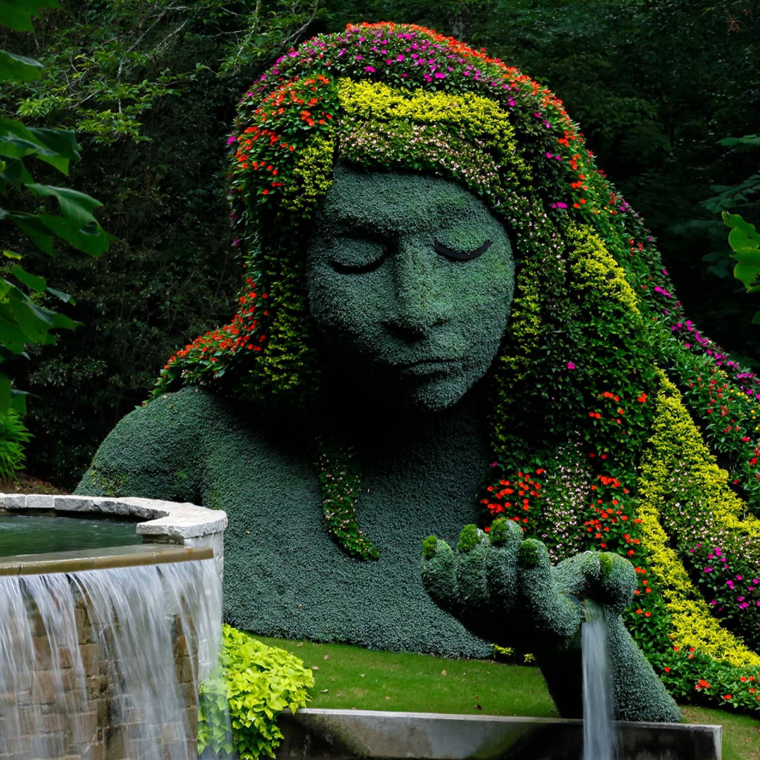 A massive statue covered in pale greenery, its hair a cascade of vibrant blooms; a fountain flows from its outstretched hand.