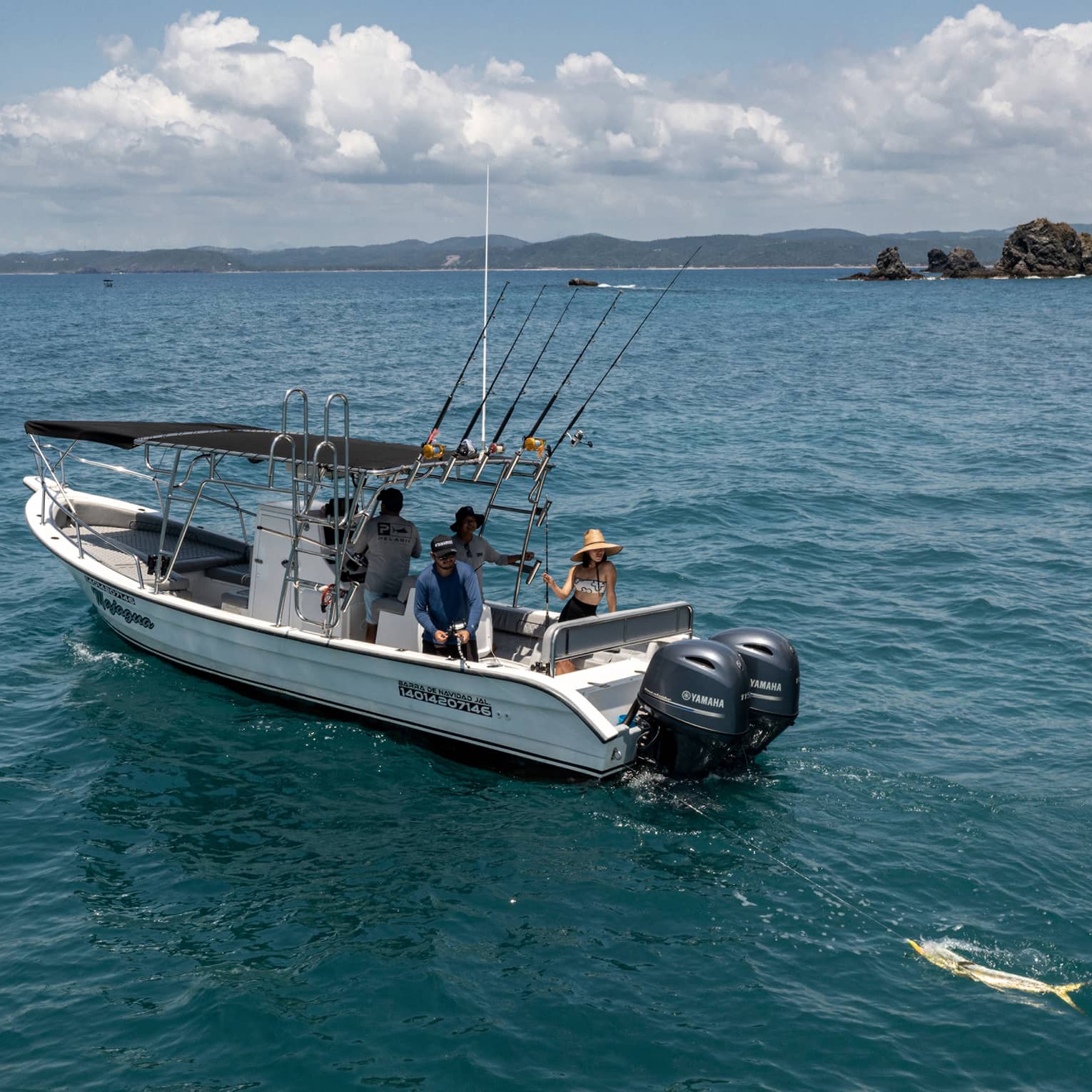 Guest reels in a large yellow fish from the back of a boat in open water near rocky islands, while two friends look on.