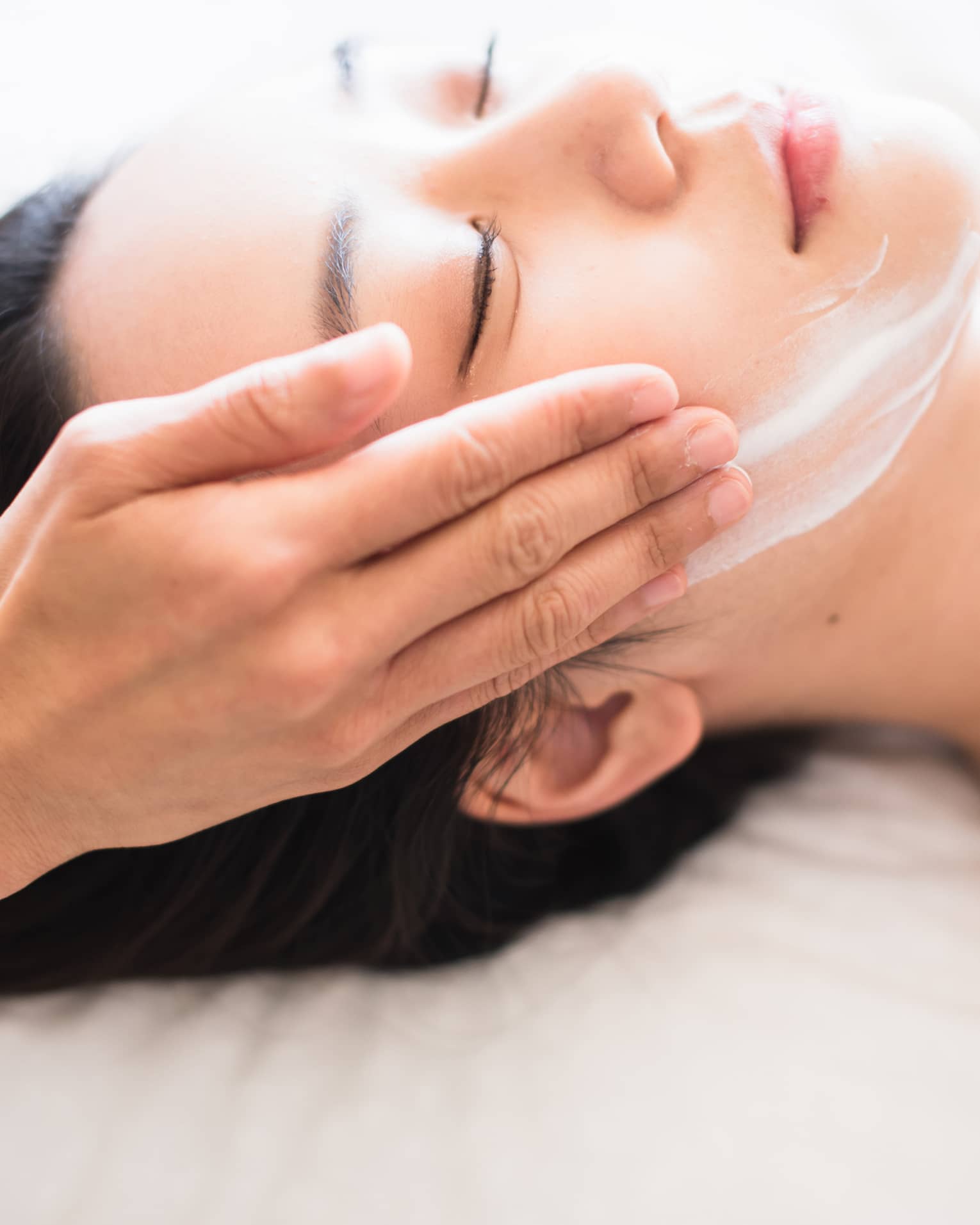 Close-up of spa attendant's hands rubbing white cream on woman's face as she lies on massage table