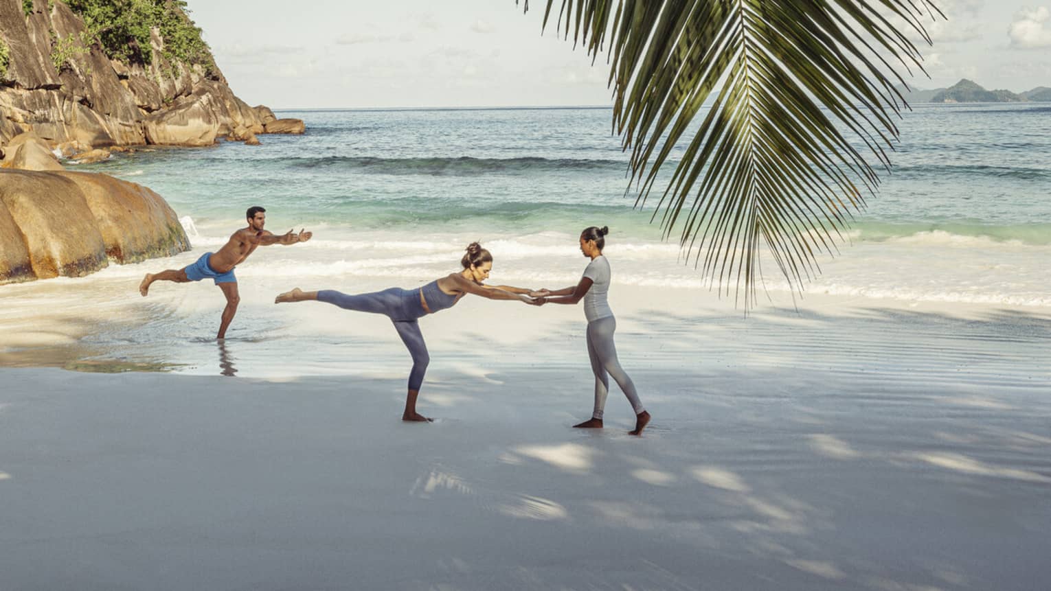 Two guests and an instructor doing yoga on a beach shaded by a large palm tree