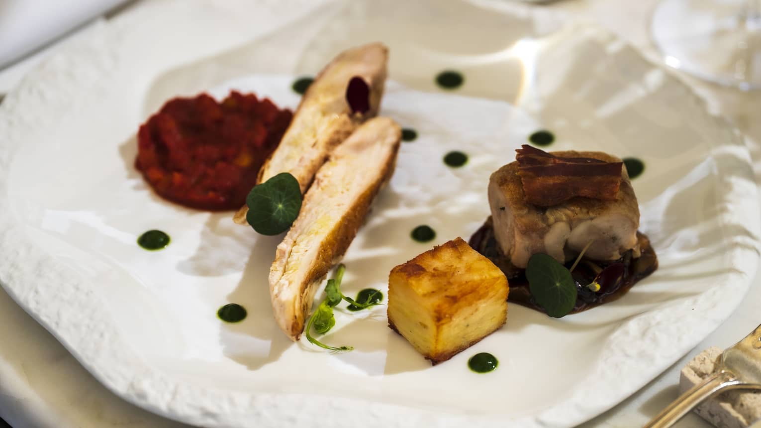 An artistic white plate with a textured rim holds food items with small dollops of sauce and garnish.