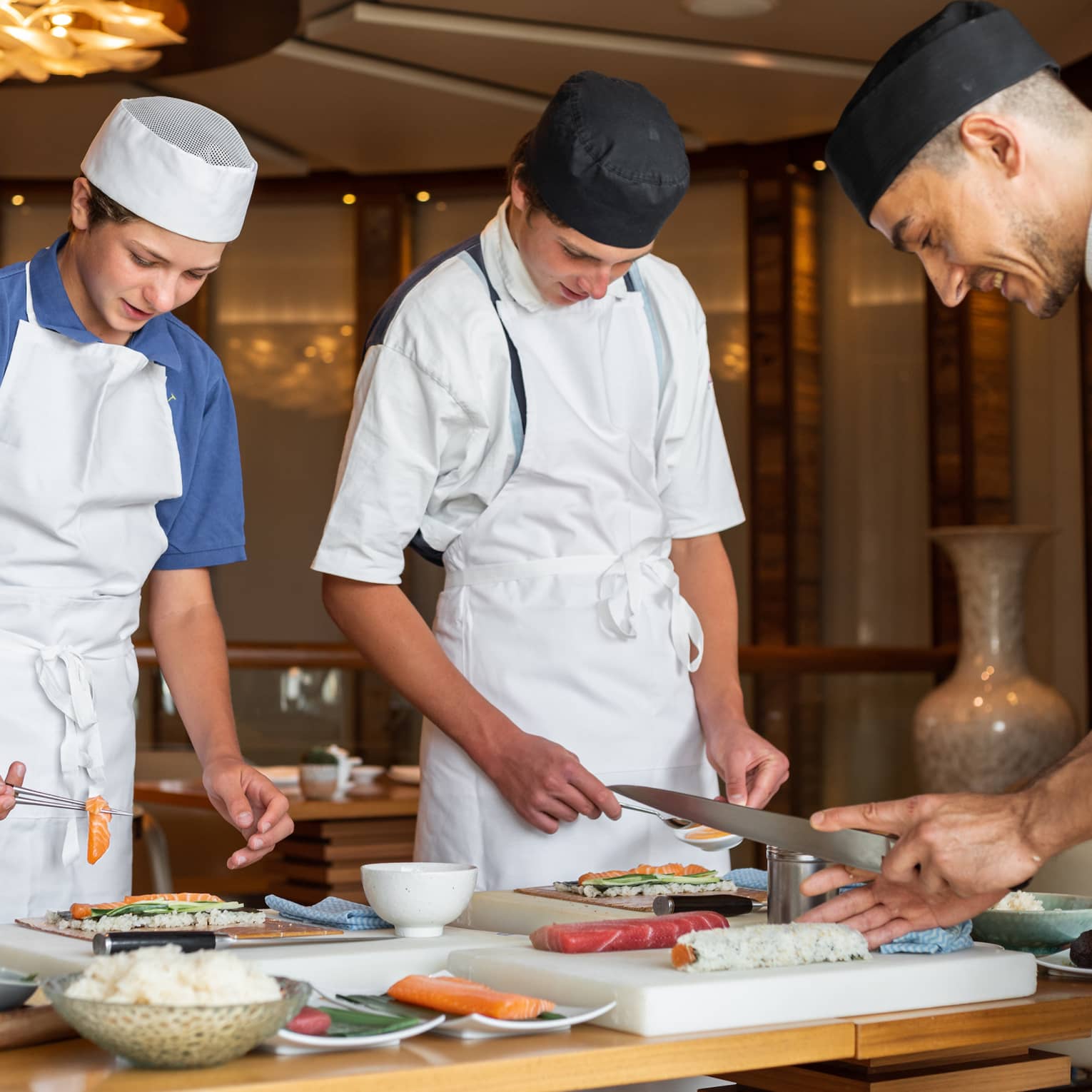 Two teenagers wearing aprons and chef hats learning to make sushi with a sushi chef