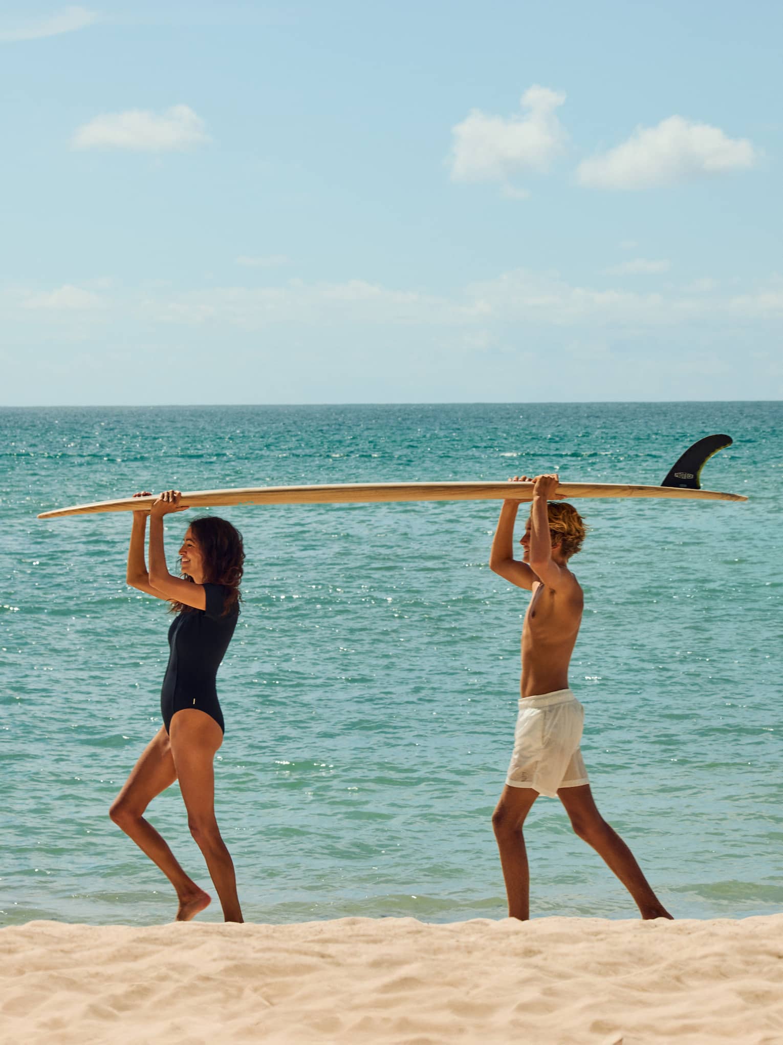 Two kids walk on beach with a surfboard on their head