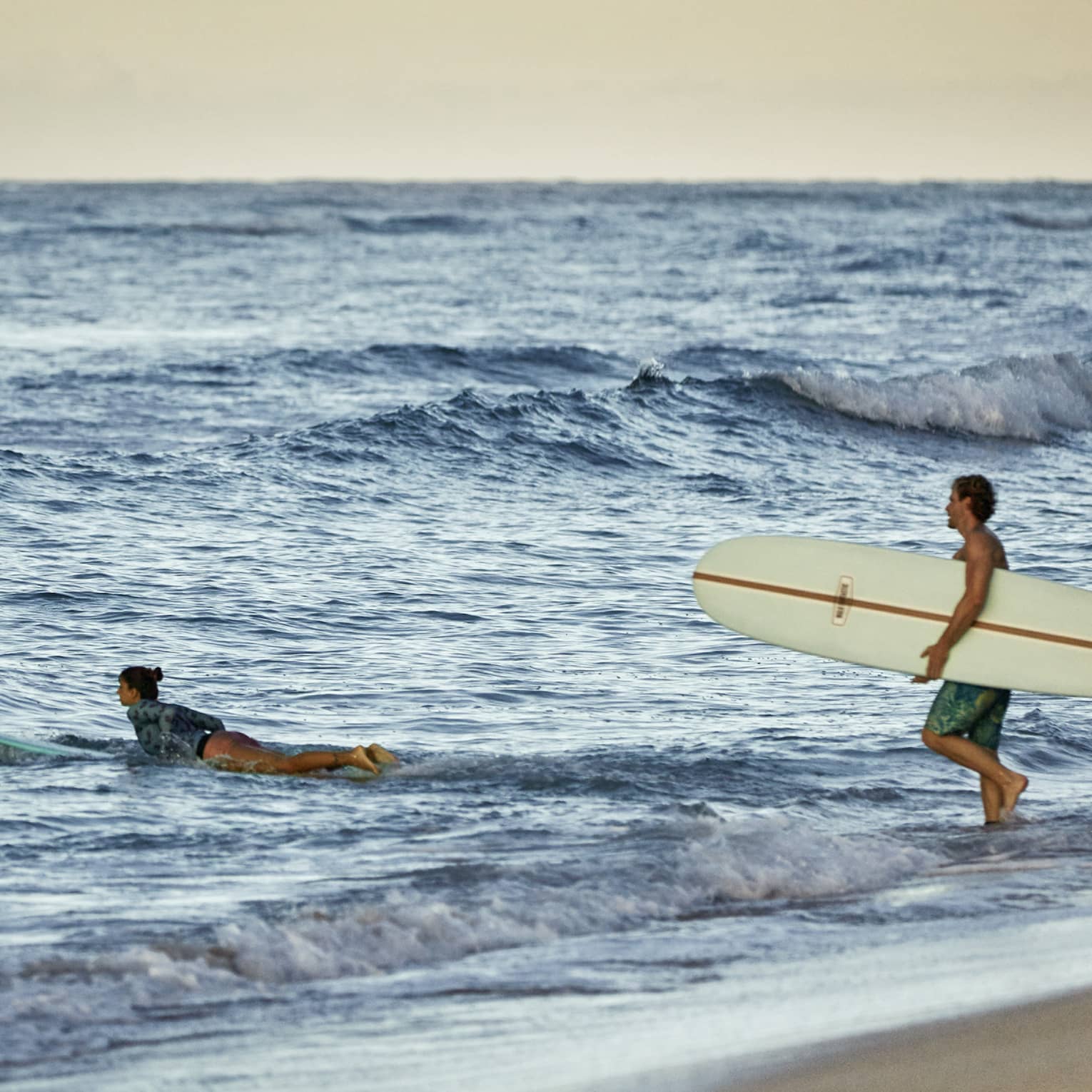 Under a golden-yellow sky at the beach, a surfer paddles their board into the waves as another wades in carrying a board.