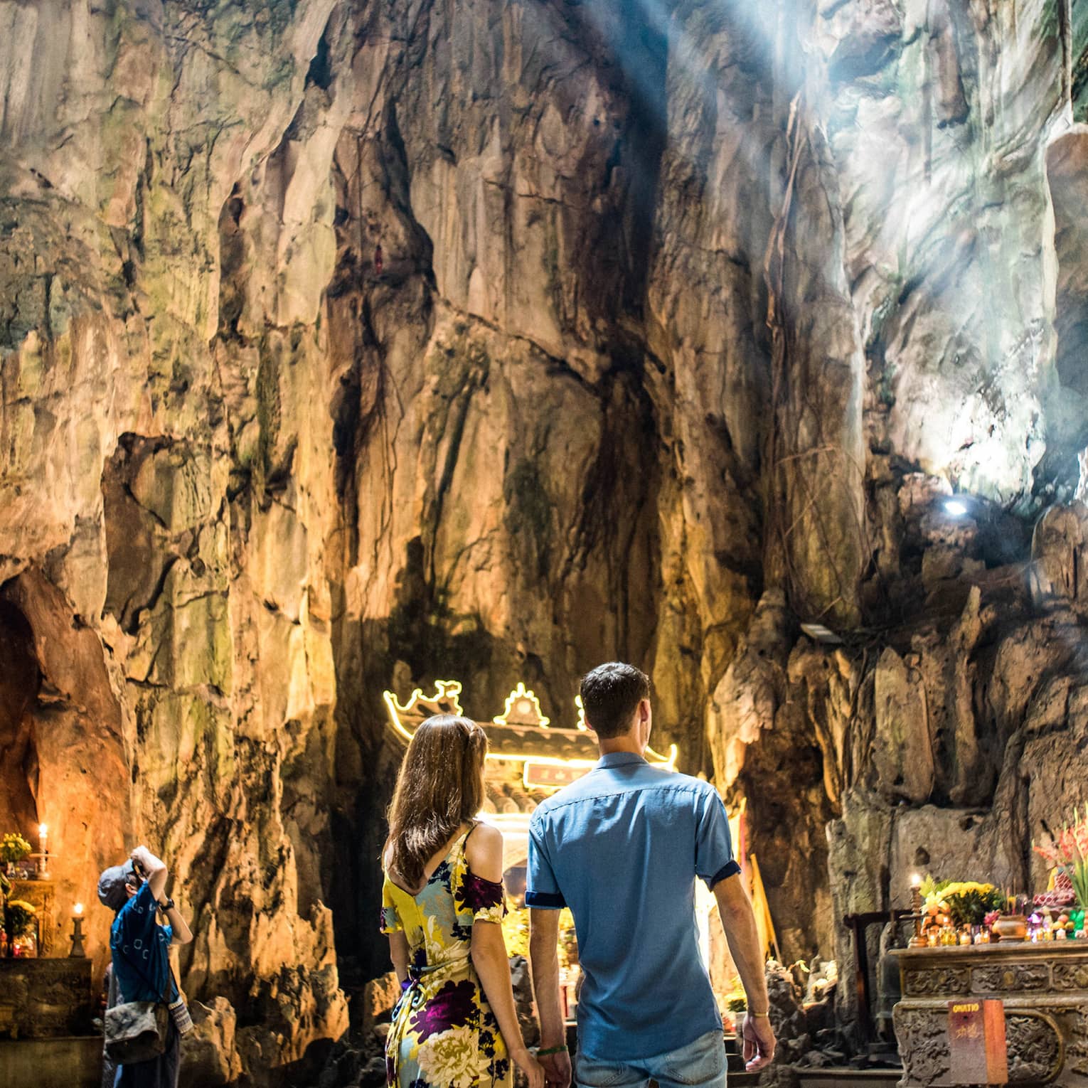 In a cave, three people look up at a large buddha statue atop a shelf in the wall; light streaming in and a shrine below.