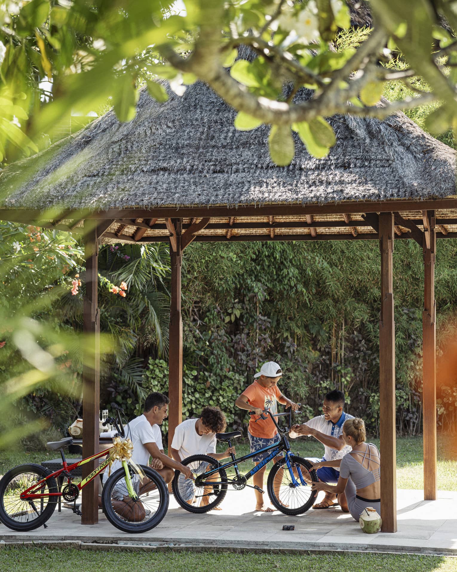 View through foliage of four adults and a child working on a bicycle under a thatched hut surrounded by lush greenery.