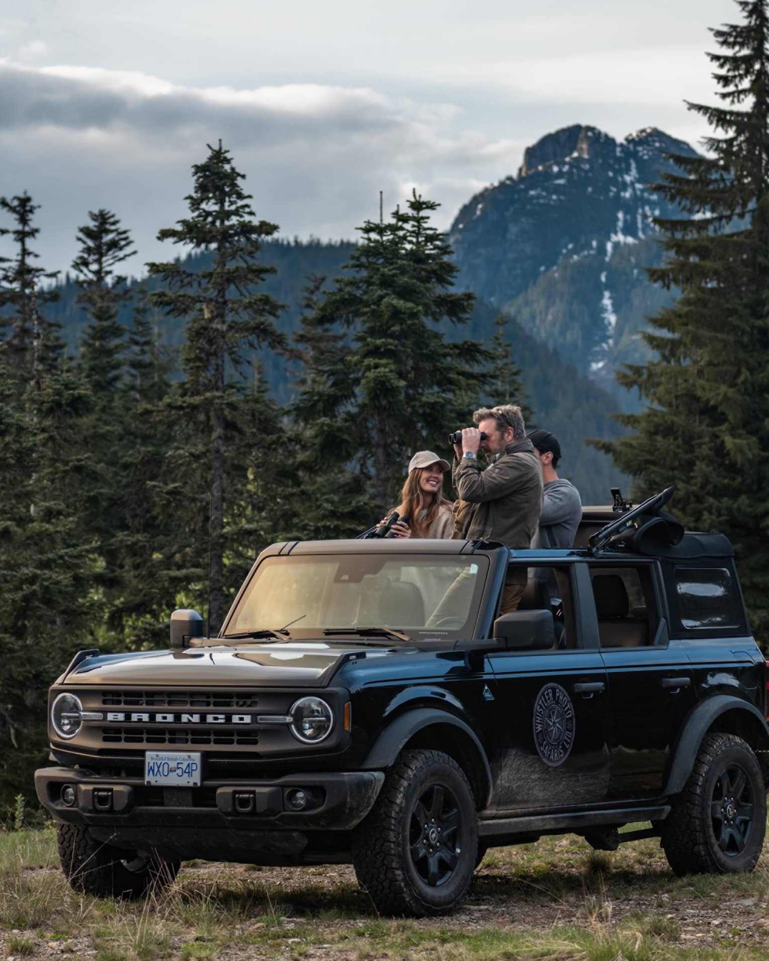 Group of friends with binoculars stand in open-top Ford Bronco surrounded by trees and mountains