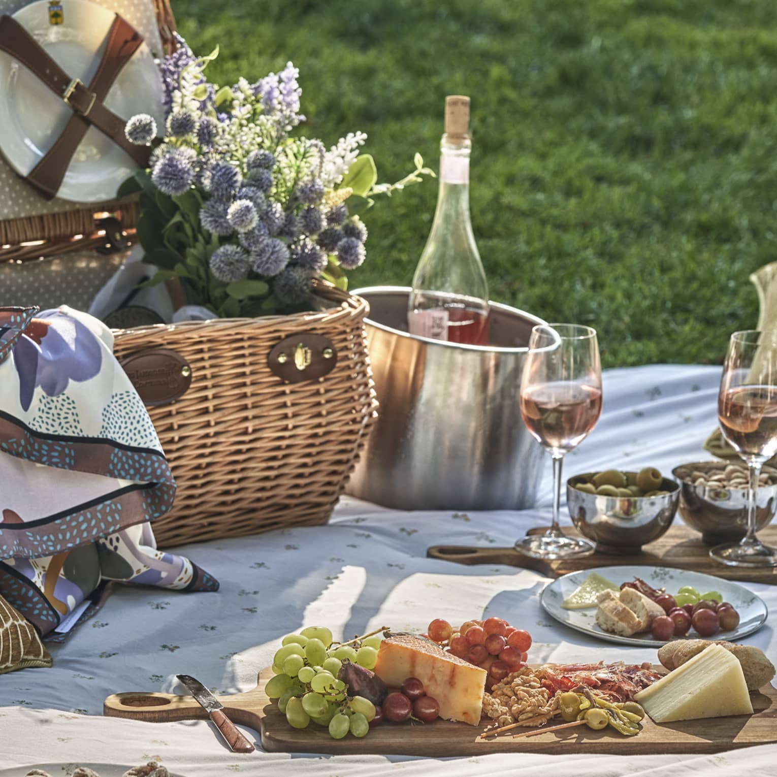 A picnic blanket spread on lush green grass is set with a fruit and cheese board, wine bucket, glasses, pillows and flowers.