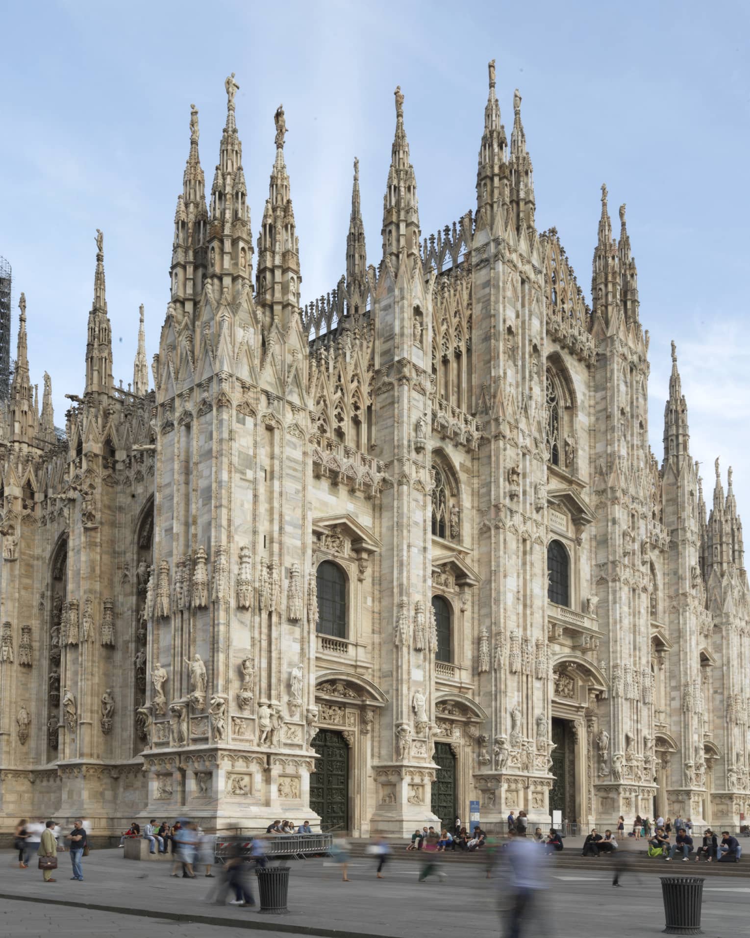 The exterior of the Milan Cathedral (Duomo di Milano), showcasing its intricate Gothic architecture with tall spires and detailed stone carvings against a clear sky.