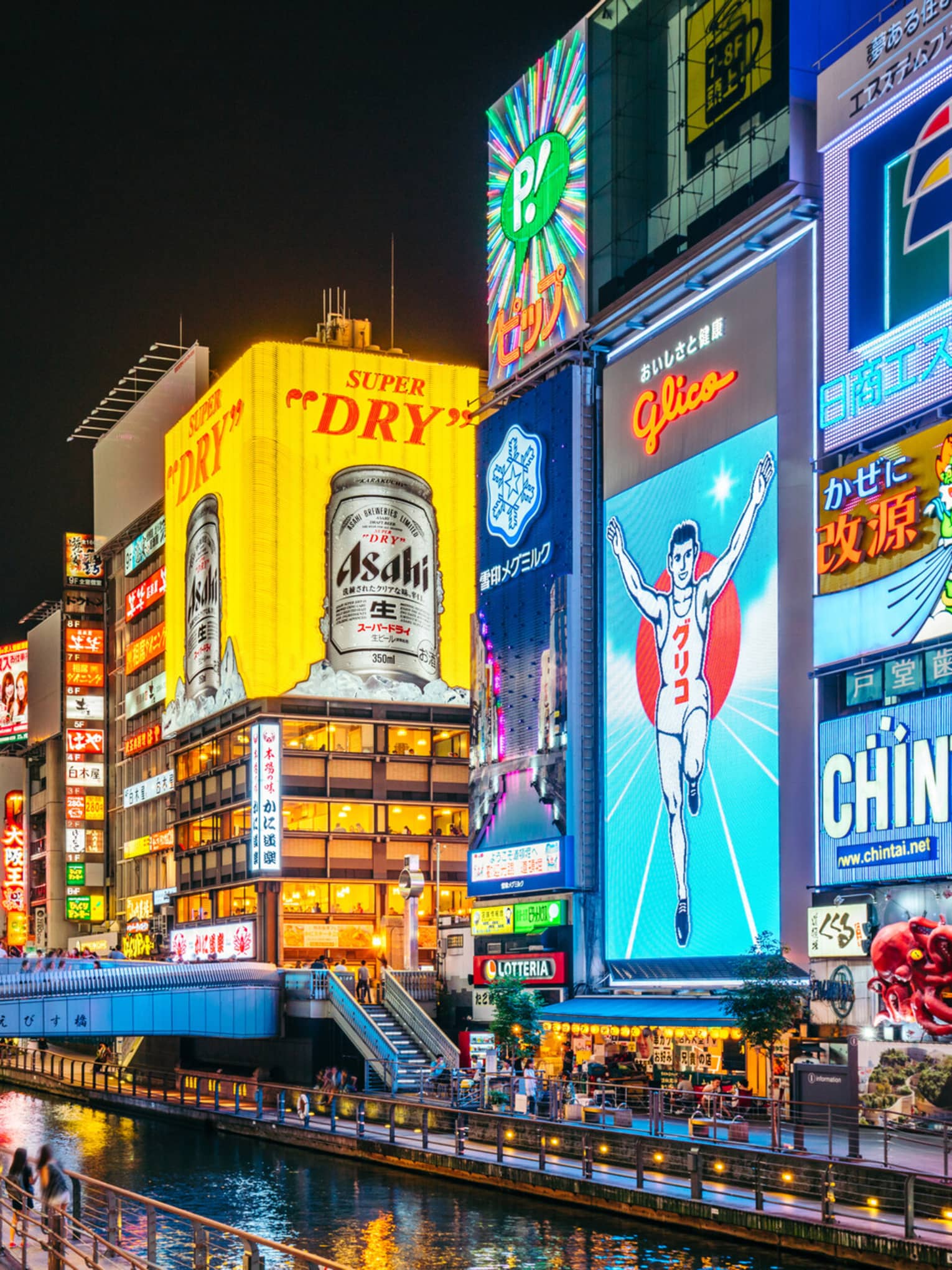 A footbridge crosses a narrow canal flanked by huge neon ads in bright colours on tall buildings in a busy city at night.