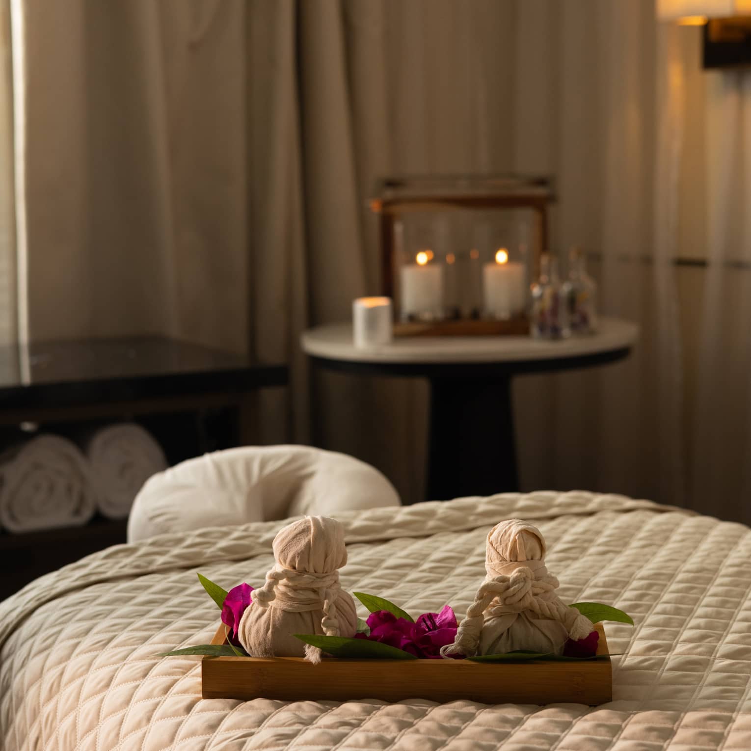 A softly lit spa treatment room withe neutral decor, with a treatment table, a window with sheer drapes admitting soft natural light, a small round table in a corner with candles and a cabinet with towels