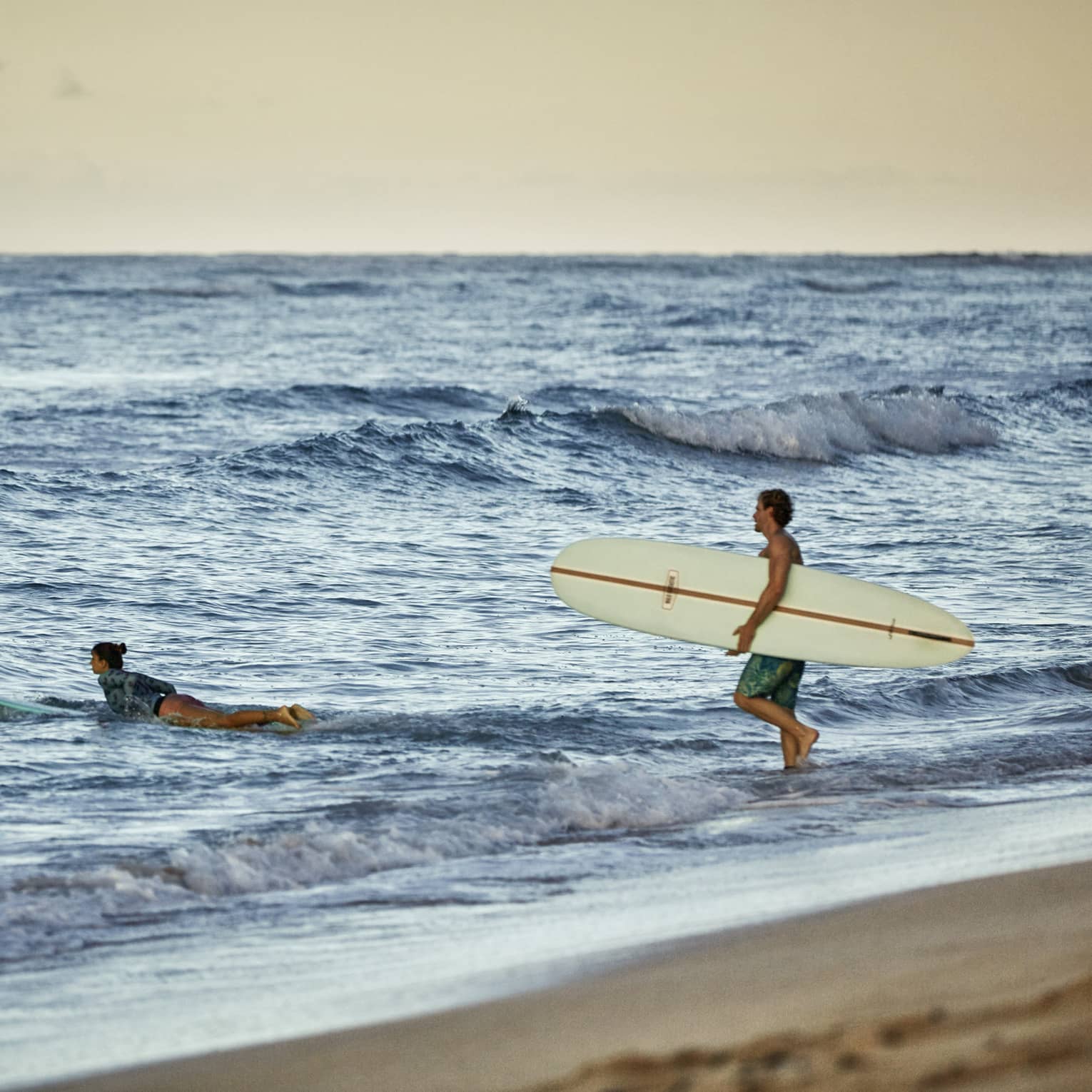 Under a golden-yellow sky at the beach, a surfer paddles their board into the waves as another wades in carrying a board.