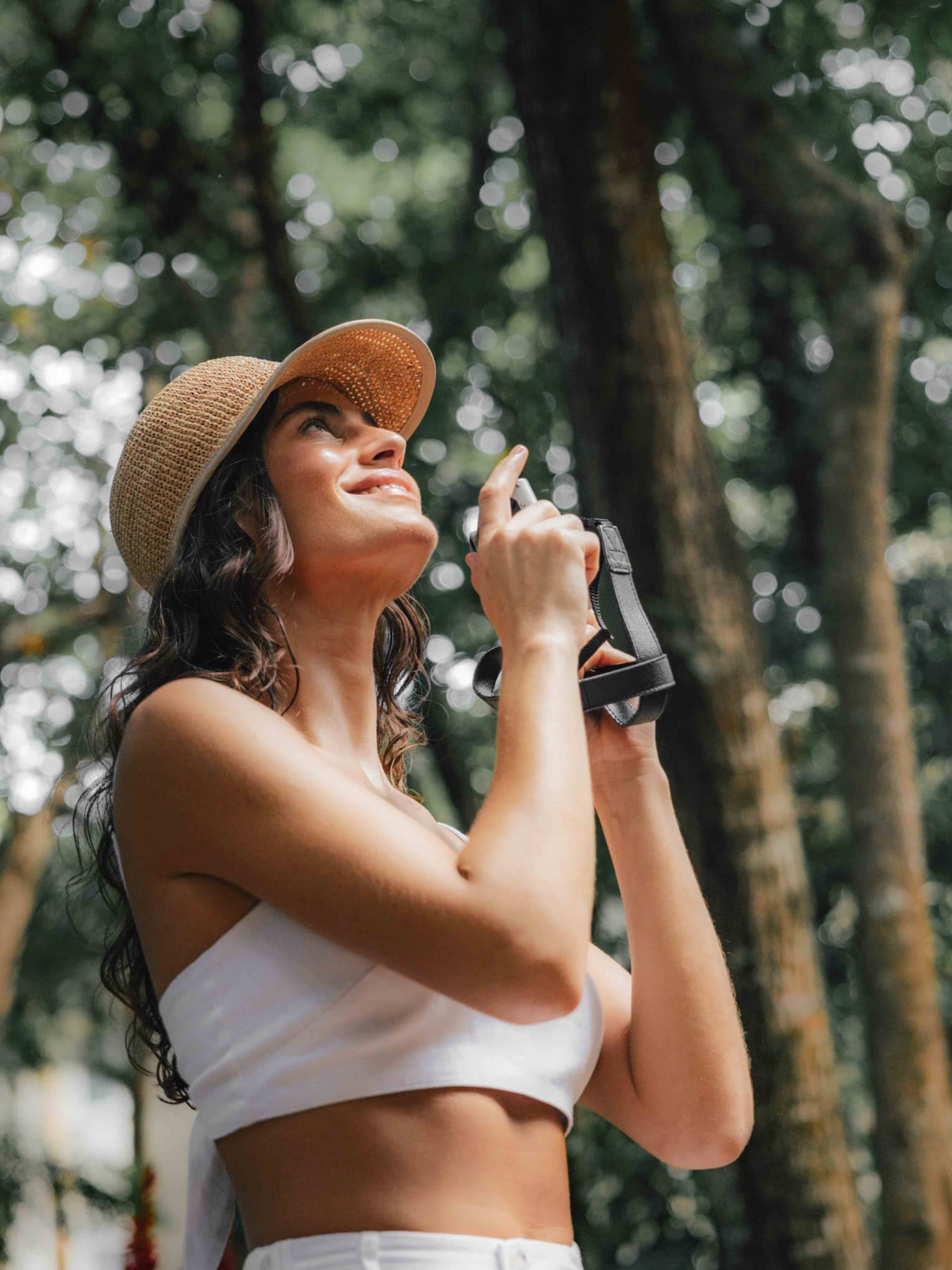 A woman stands in a tropical forest holding a camera, looking up