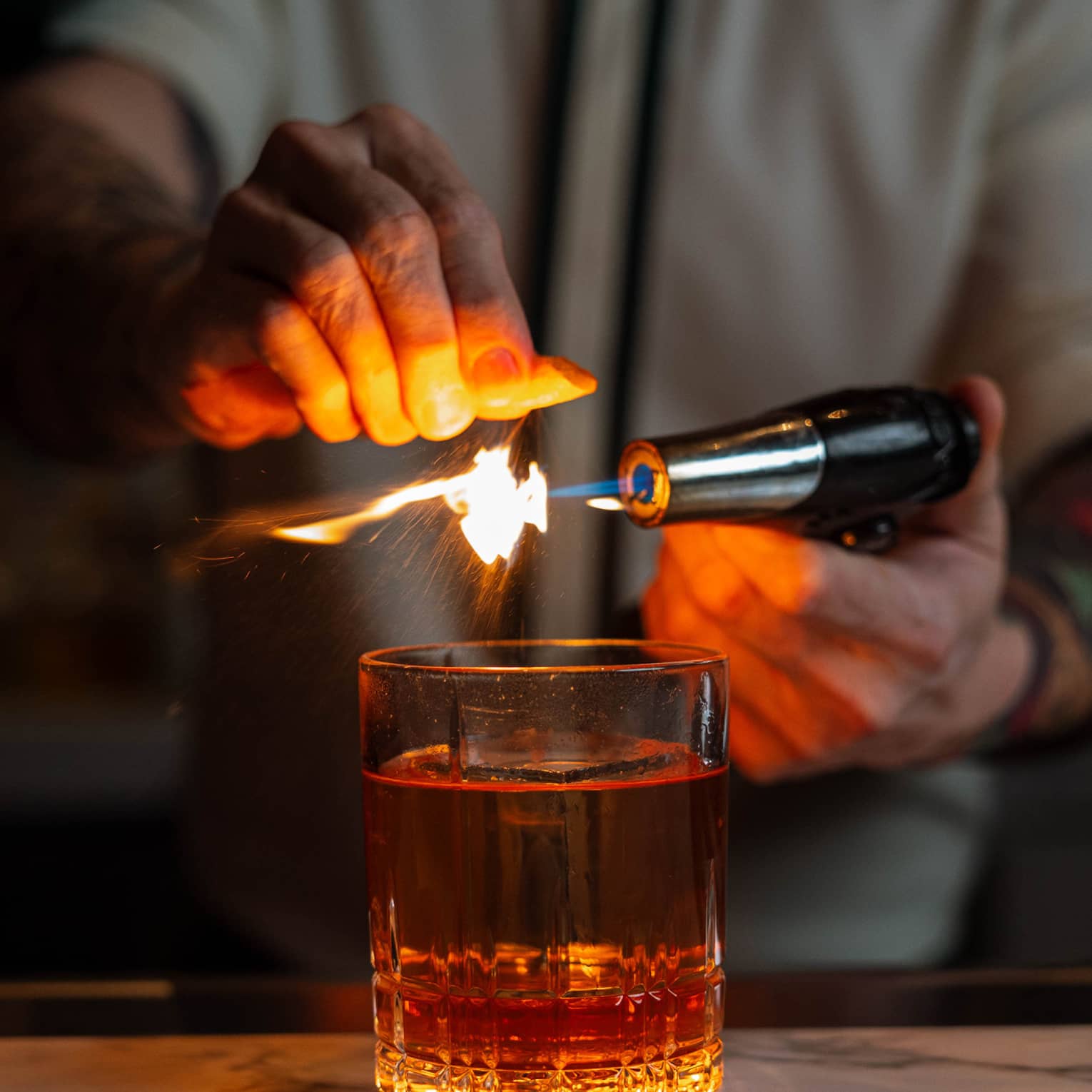 Bartender using a torch to flame an orange peel over a glass of whiskey, creating a dramatic effect at the bar.