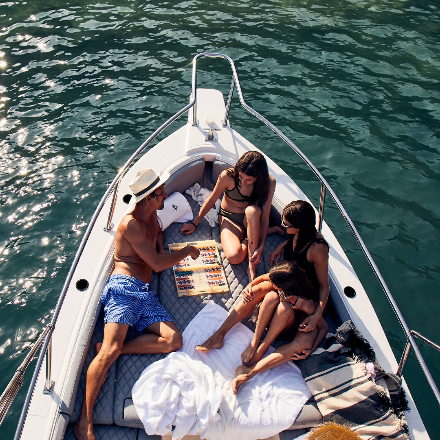 A family lounges in the bow of a luxurious boat amid towels and blankets, playing backgammon, the sun's glow on teal waters.