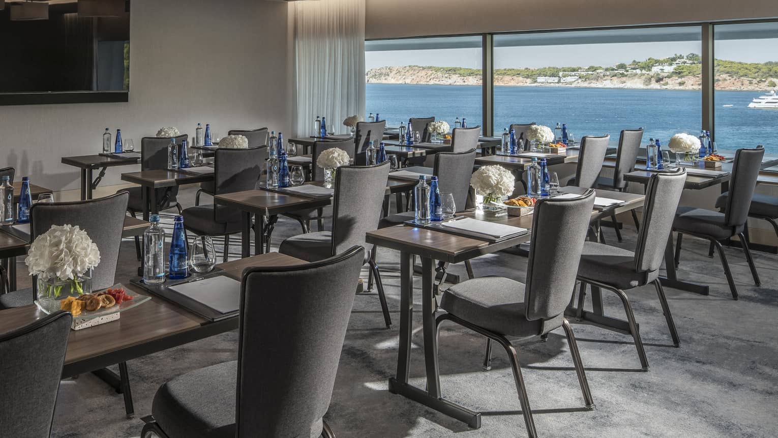 Rows of desks with water, flowers, snacks and grey chairs facing TV with ocean view
