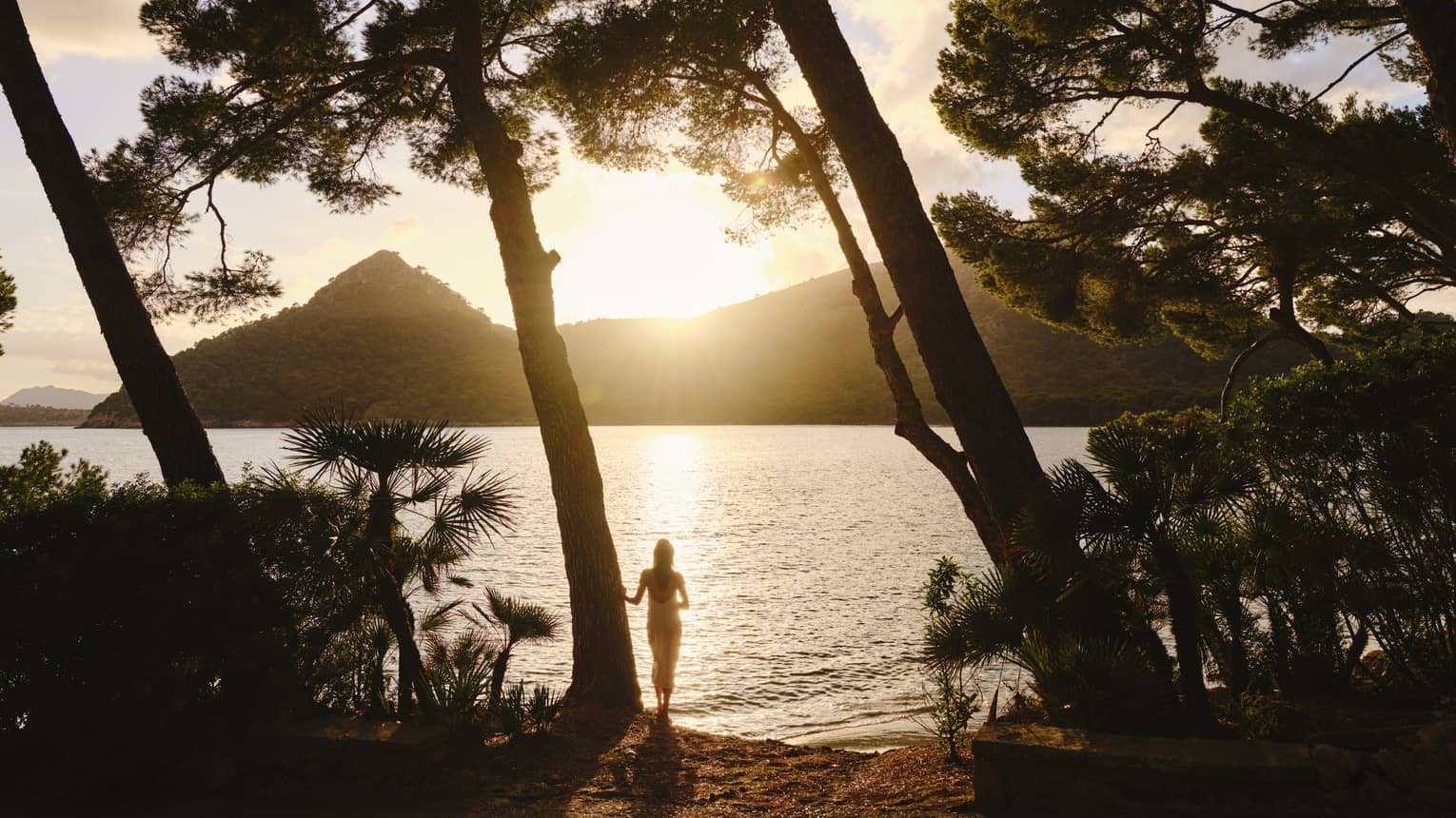 Silhouetted by the sun dipping behind a mountain, a guest stands at the edge of rippling water framed by tall leaning trees.