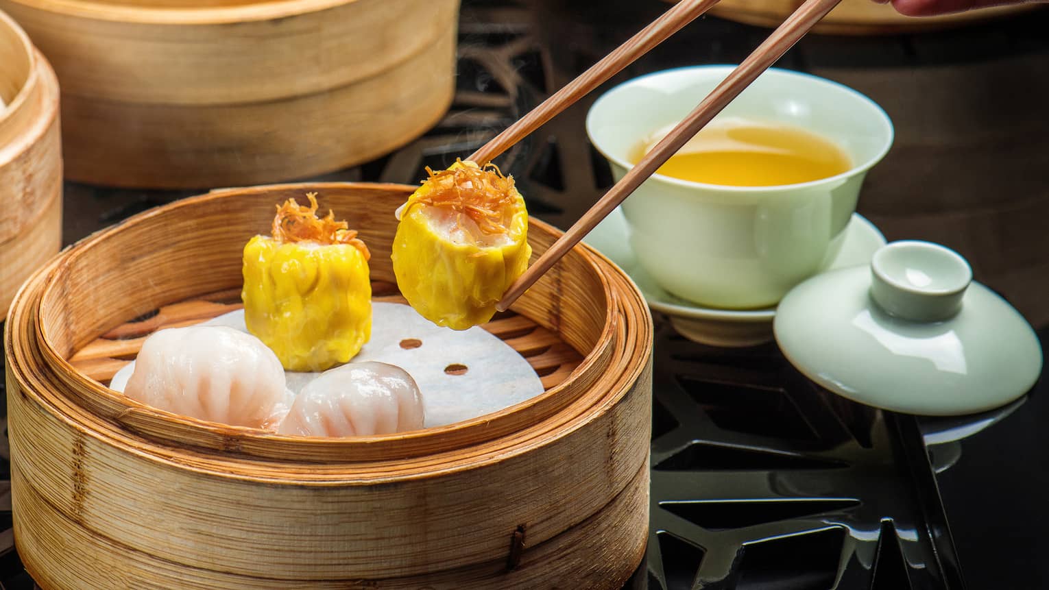 Close-up of hand holding chopsticks, dim sum dumpling over bamboo steam baskets 