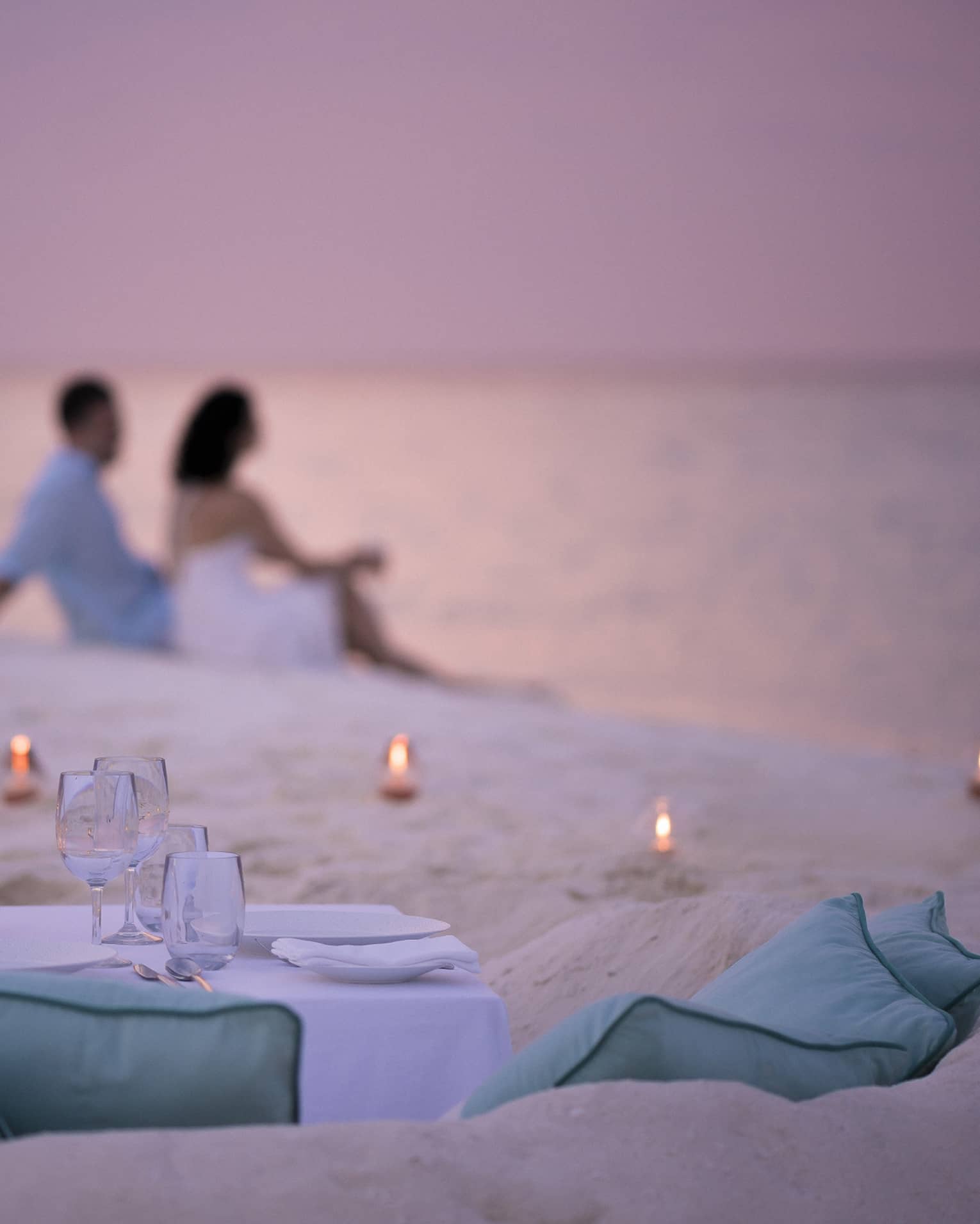 Private dining table in front of sand beach with tealight candles, couple sitting in sand