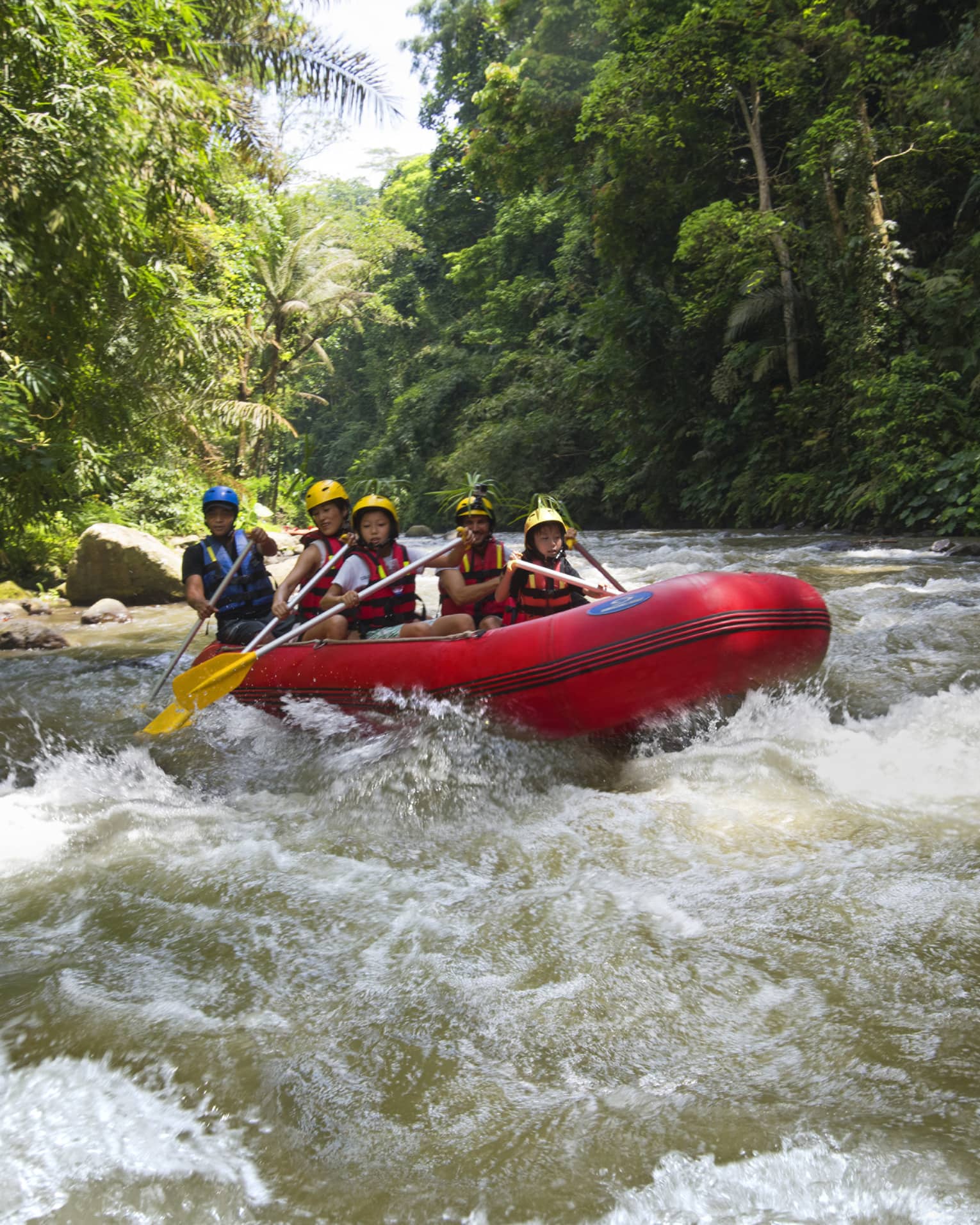 Family of four and guide hold paddles, steer red inflatable raft on rapids in river