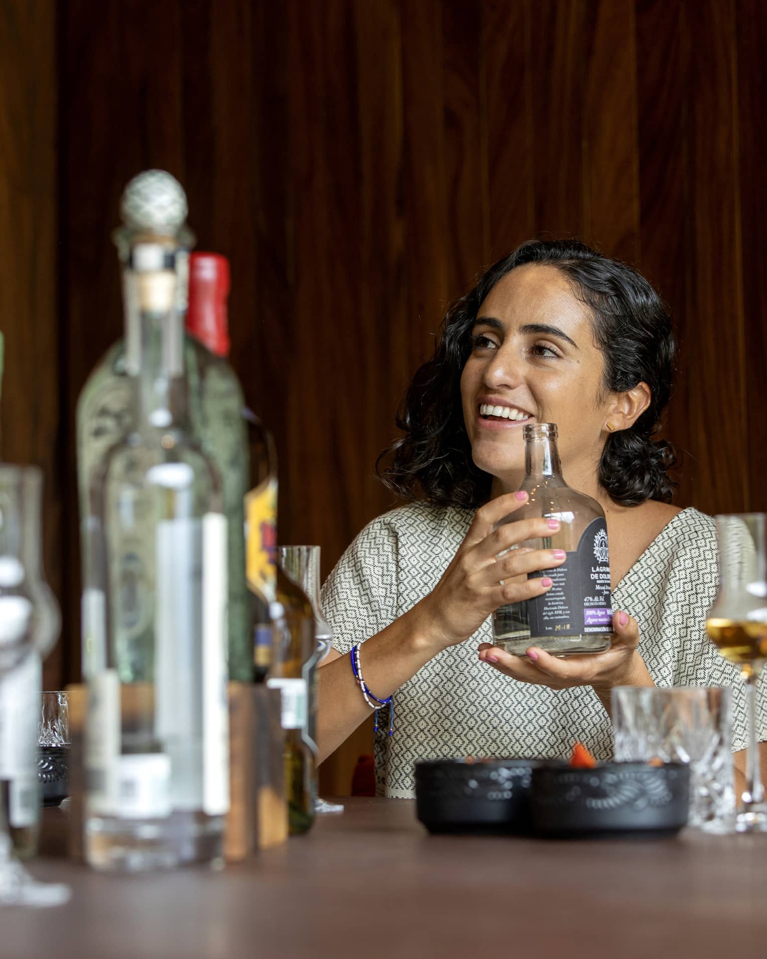 A guest cradles a short, wide bottle of liquor at a table littered with bottles of various sizes and grappa glasses. 