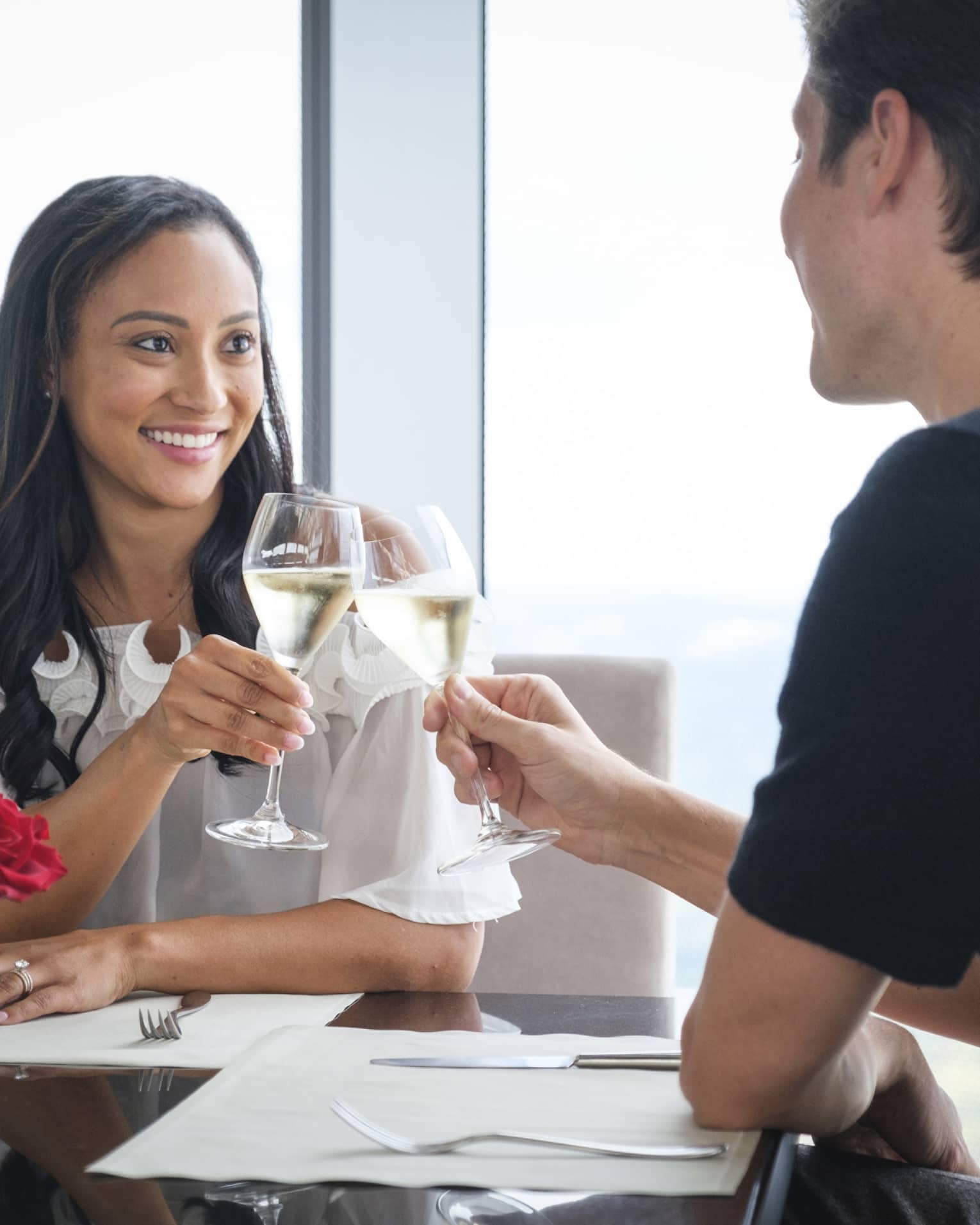A man and woman celebrating with wine and red roses on a table near a large window.