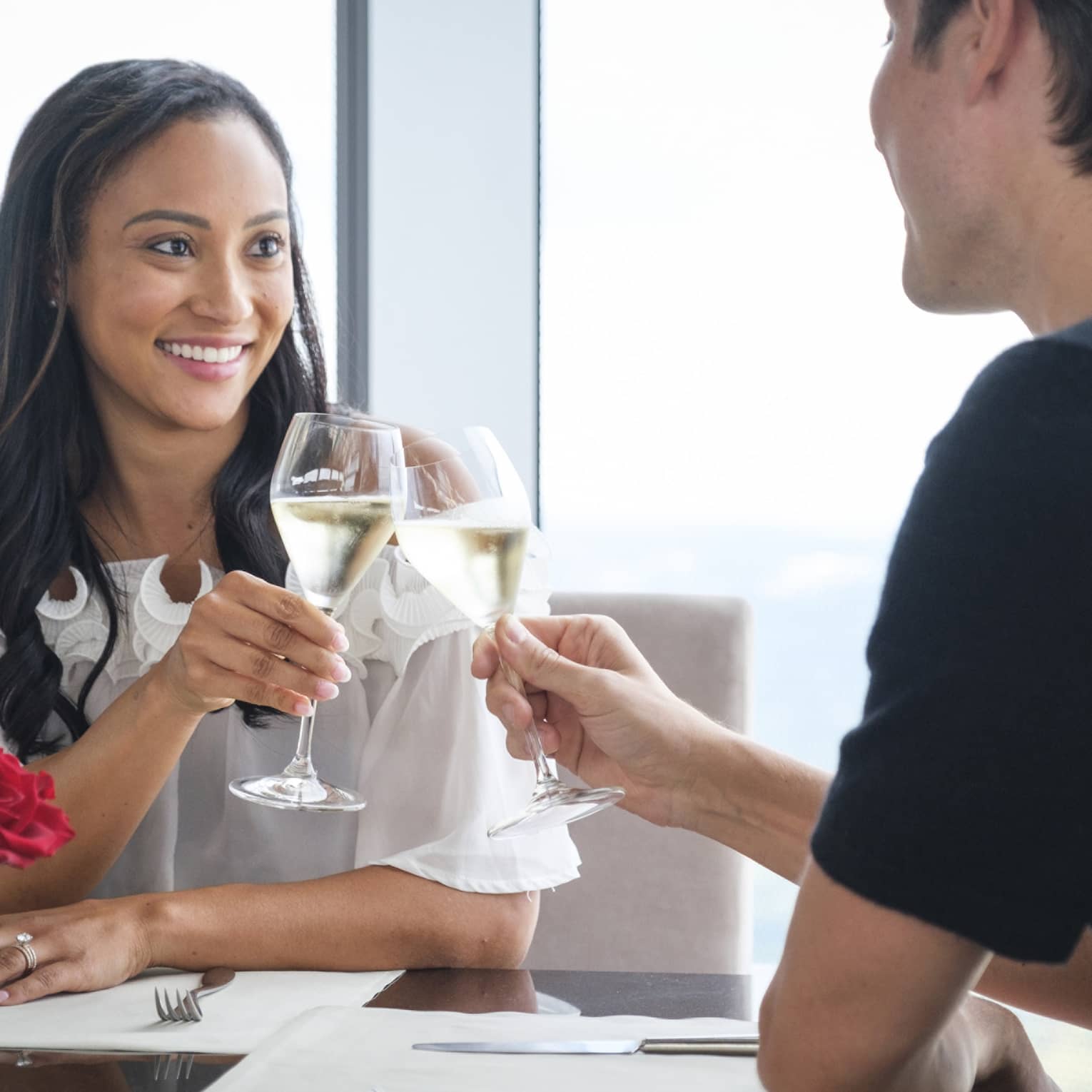 A man and woman celebrating with wine and red roses on a table near a large window.