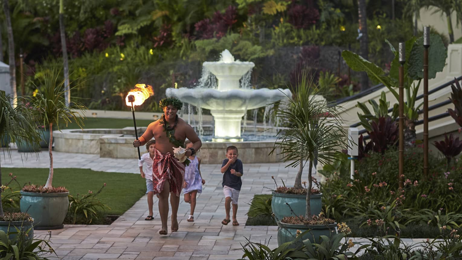 Three laughing children follow a Hawaiian torchbearer as he runs on a stone path in a lush tropical garden at dusk.
