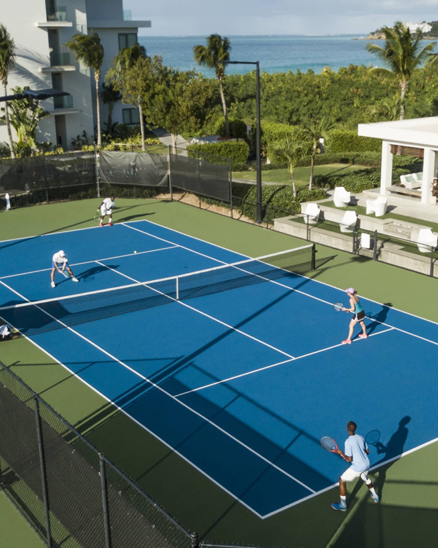 Aerial view of a doubles match on a blue tennis court, with green bordering, palm trees