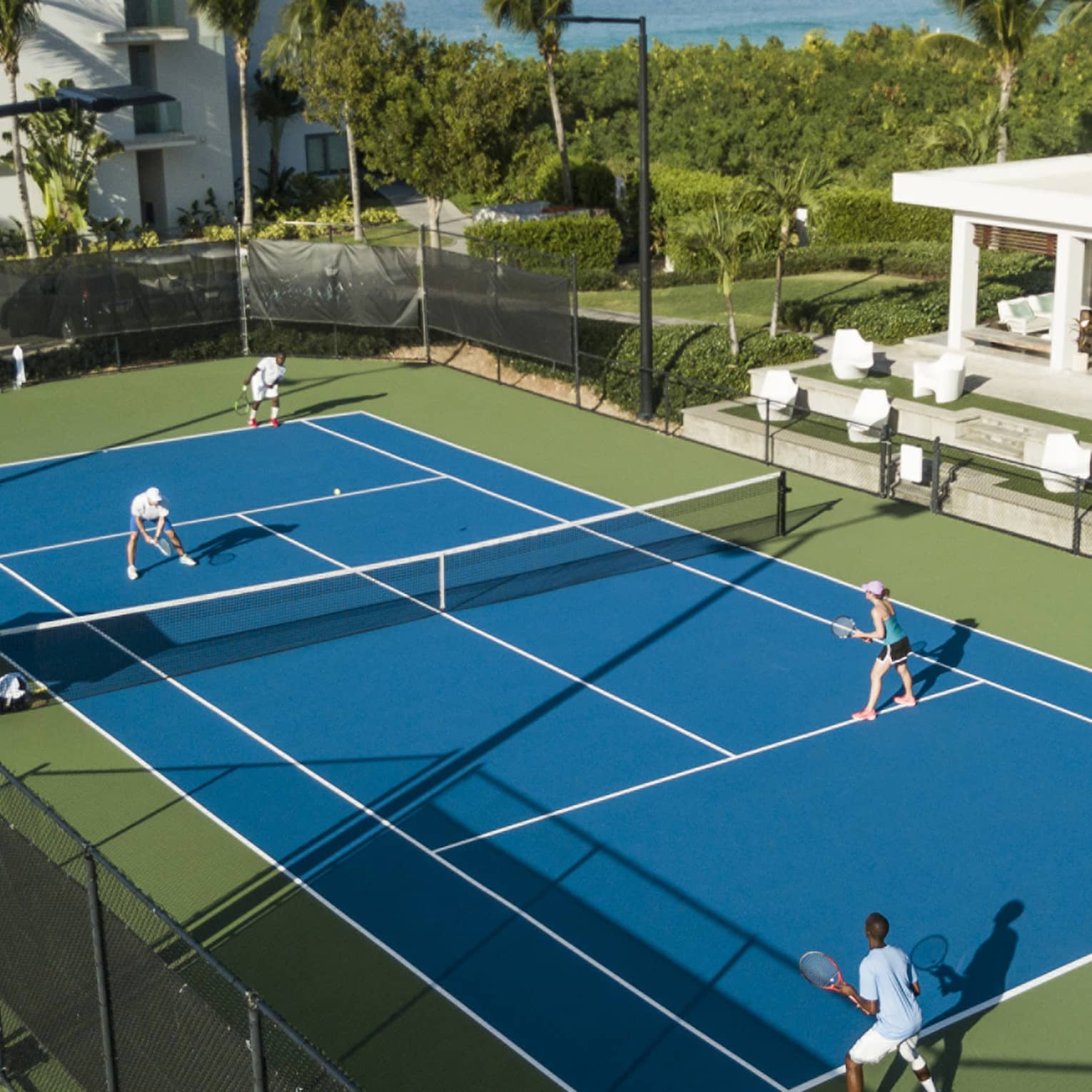 Aerial view of a doubles match on a blue tennis court, with green bordering, palm trees
