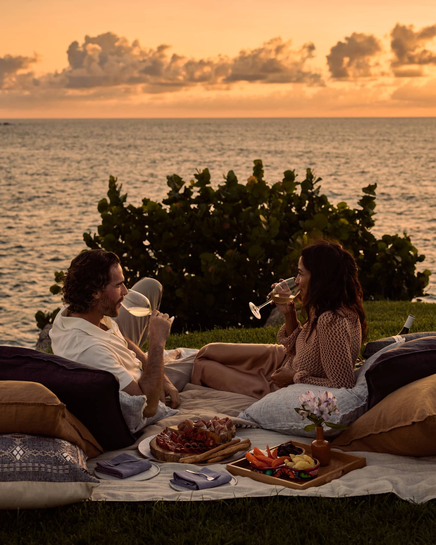 A man and woman lay on blanket and pillows during luxury picnic at sunset, next to the ocean