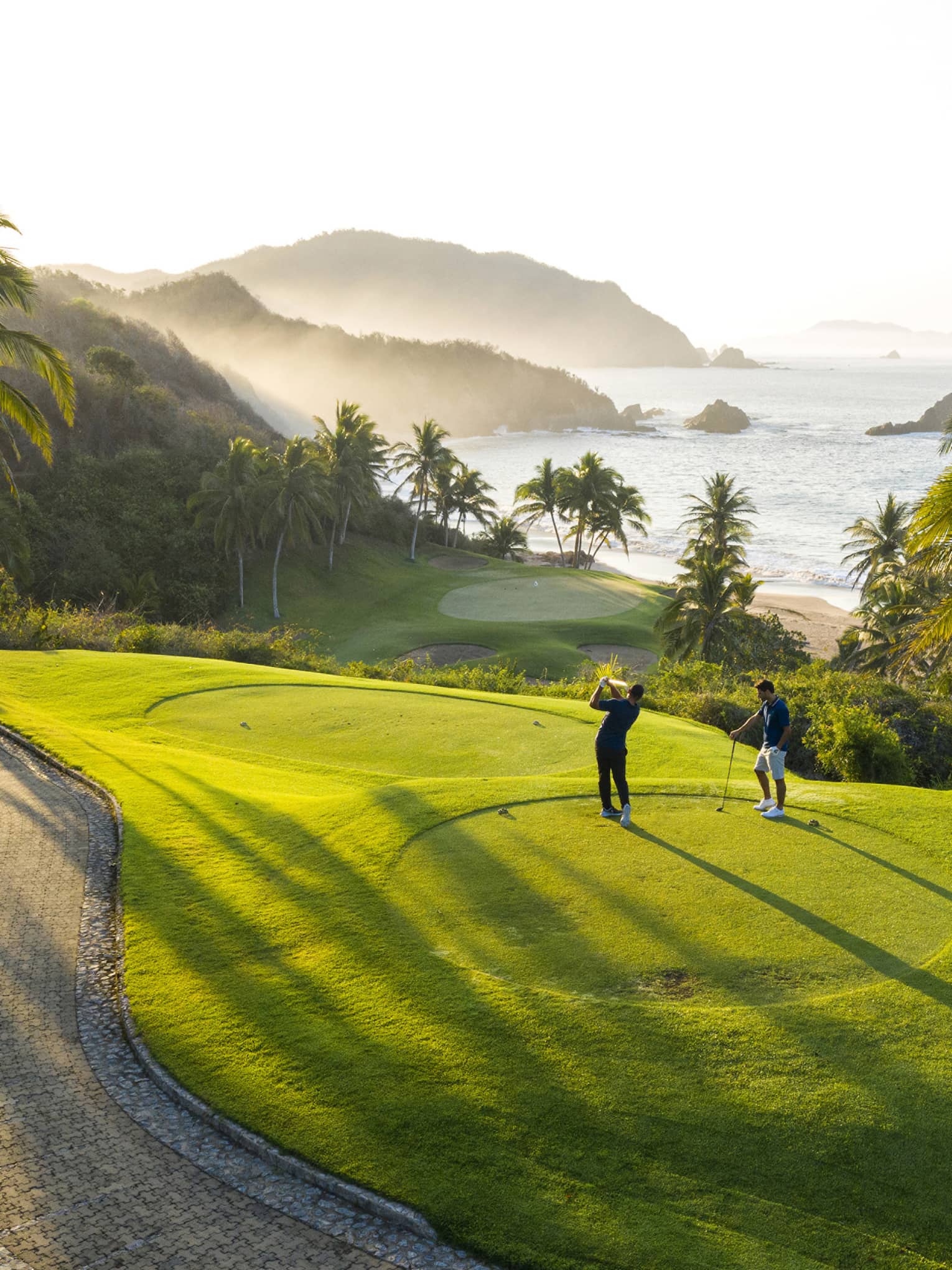Rear view of two golfers on an elevated tee, one swinging toward the green down by the beach, misty hills and ocean beyond.