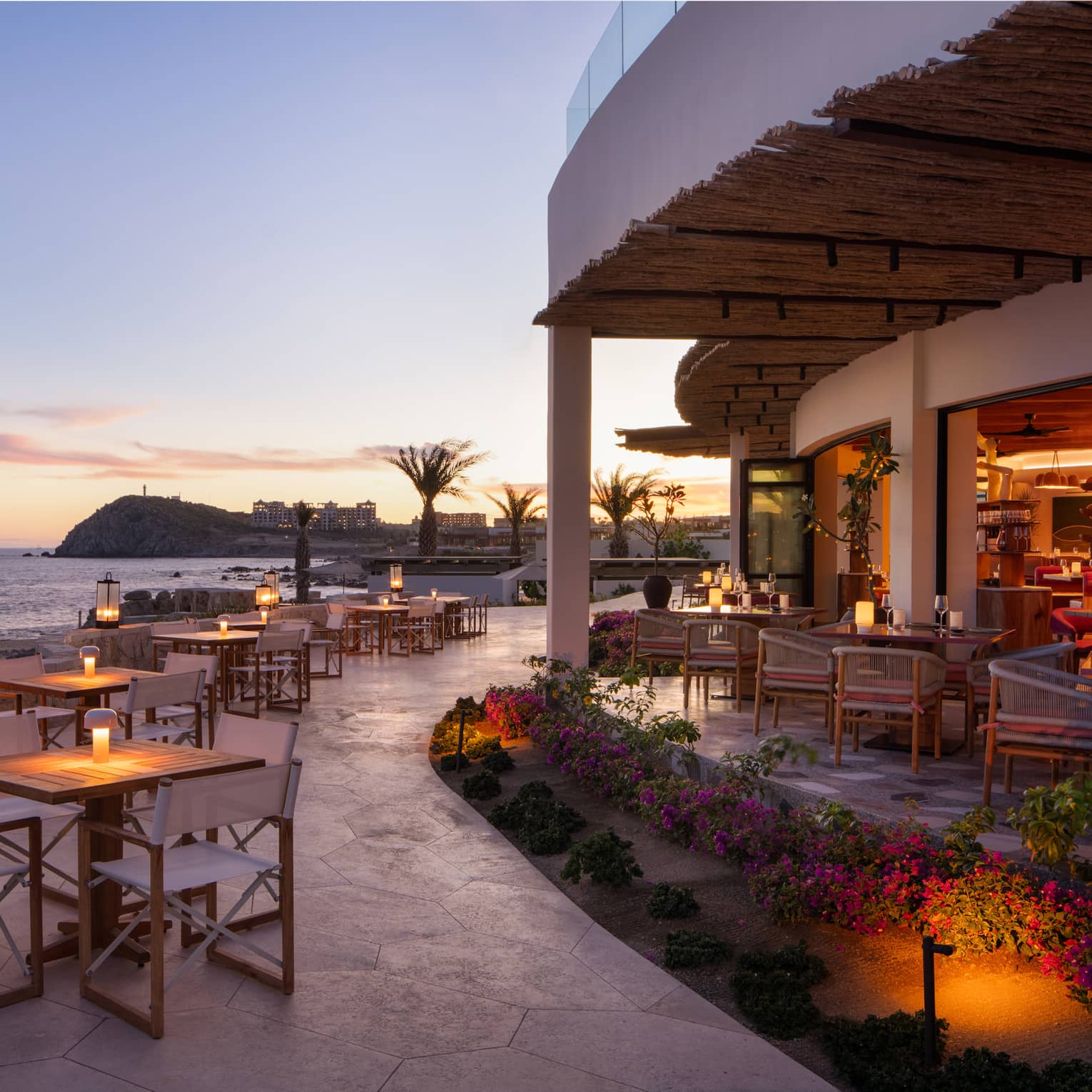 In the warm afterglow of sunset, lanterns light up a large, curved stone restaurant patio at the edge of the ocean.
