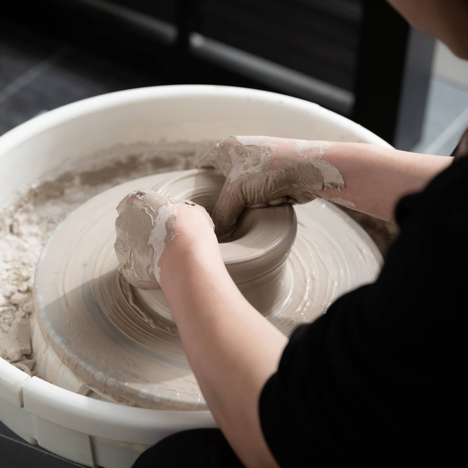 Close-up of two hands covered in wet beige clay shaping a pot on a spinning pottery wheel. The potter is wearing black.