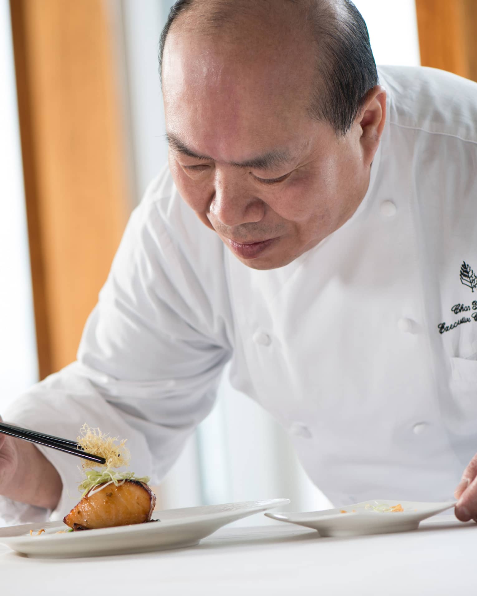 Chef in white uniform leans over grilled salmon dish on plate, adds garnish with chopsticks