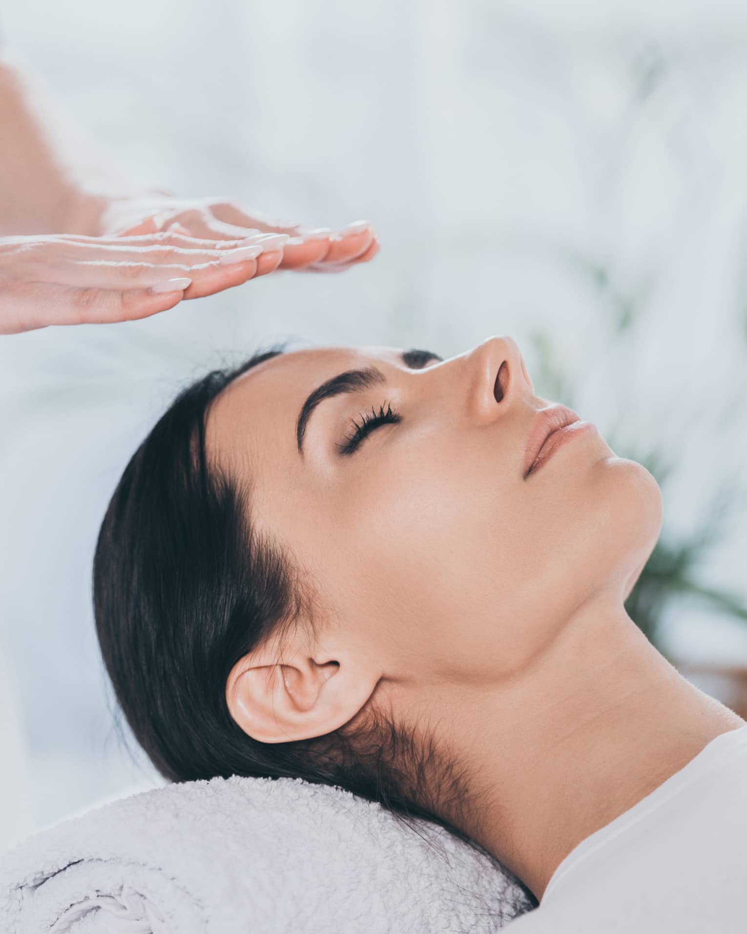Woman with eyes closed lays on rolled up towel on spa table under therapists' hovering hands