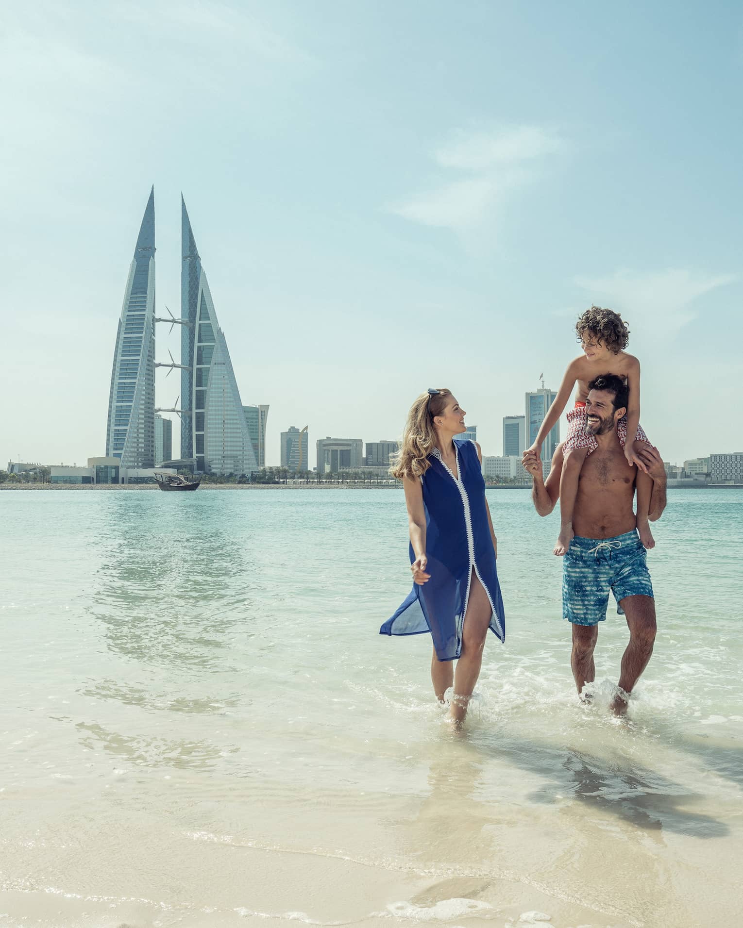 A man with a teenager on his shoulders walks with a woman through shallow ocean water with the city of Bahrain Bay in the background 