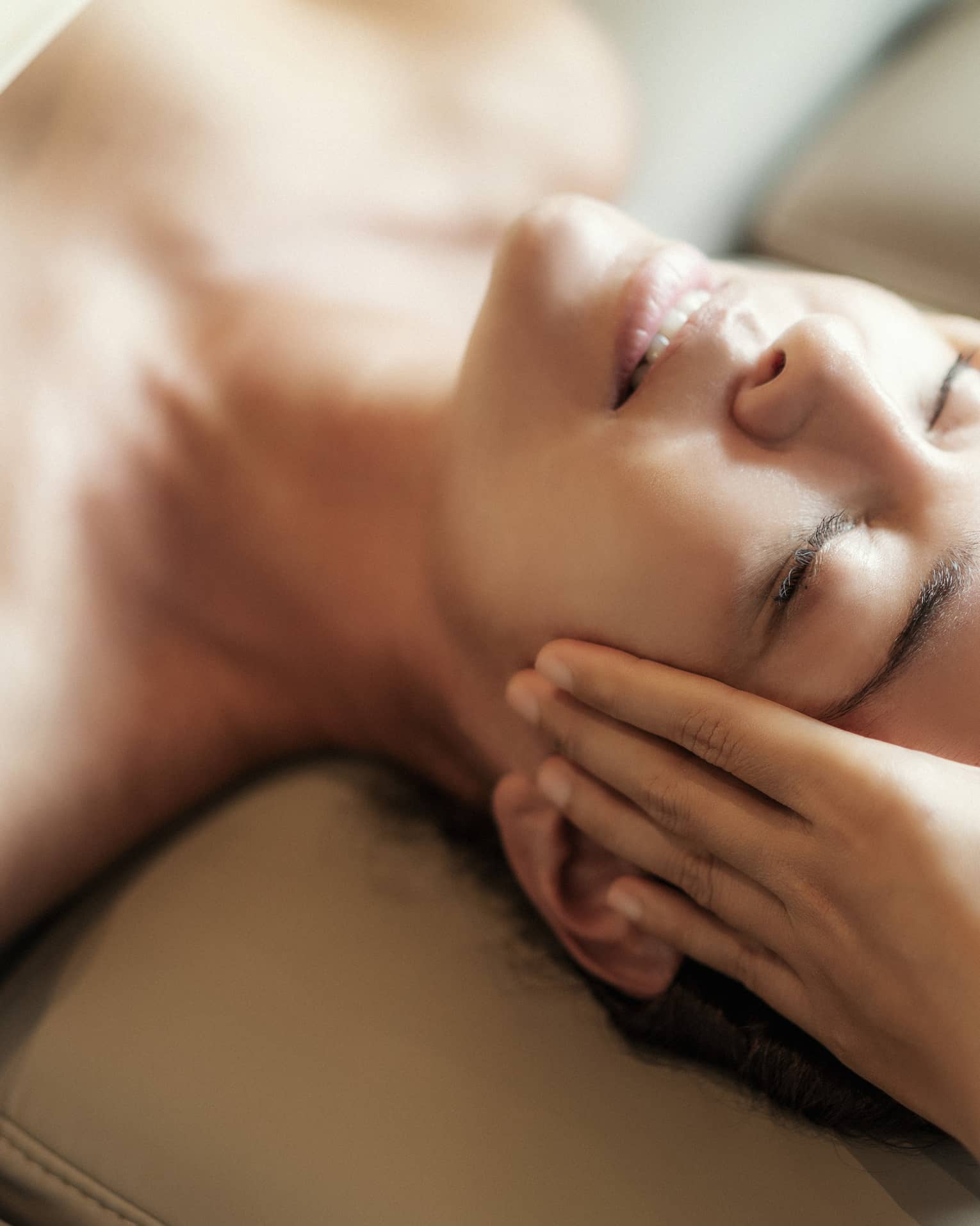 Spa facial, two hands rest on woman's forehead as she closes her eyes, lays under sheet on treatment table