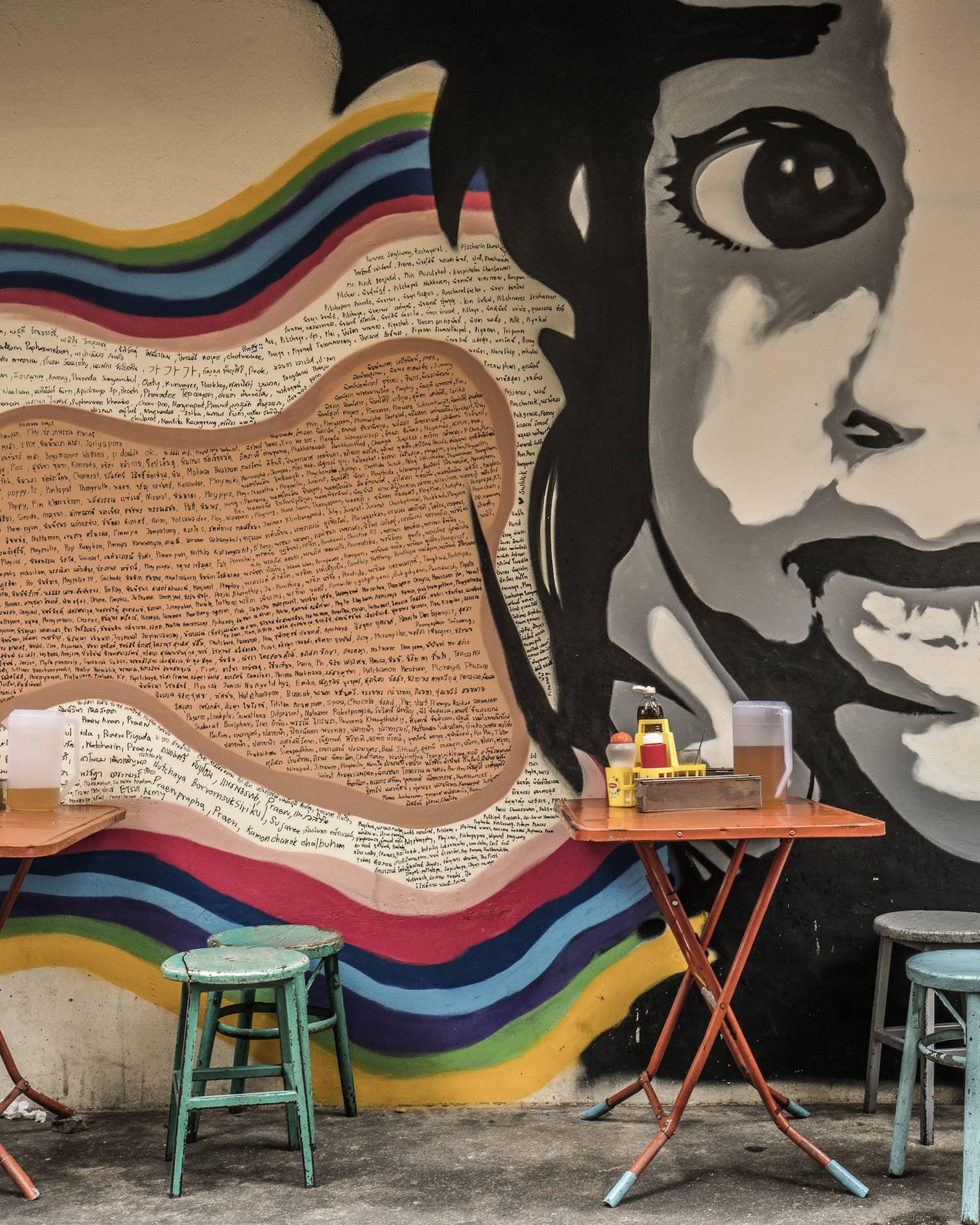Street art mural featuring a large black-and-white face on a wall, with small, colourful dining tables and stools in front it