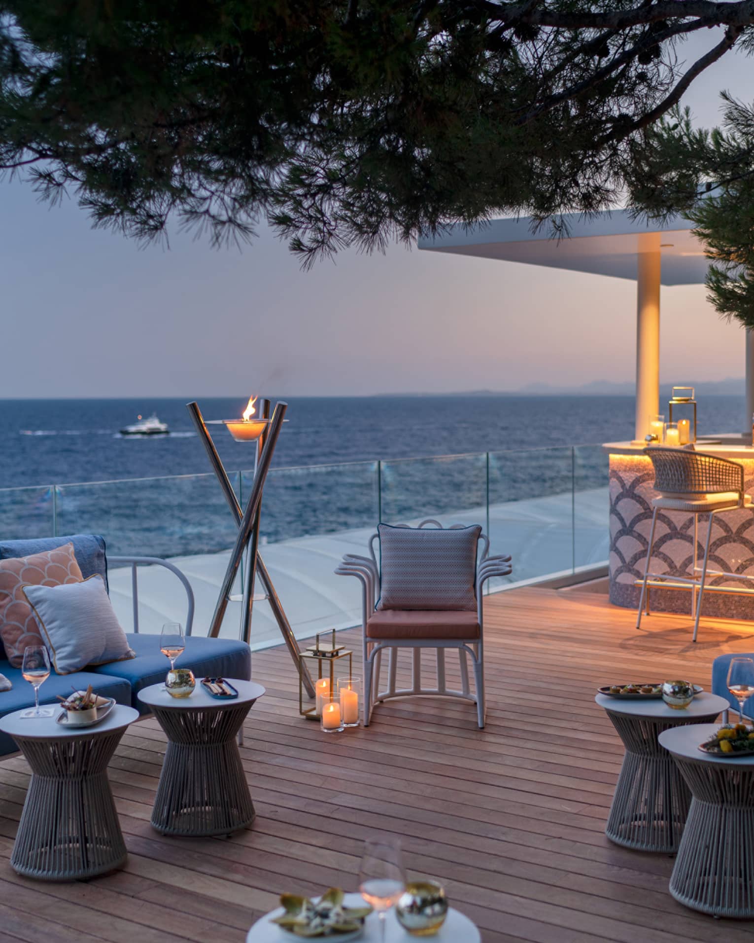 Club Dauphin wooden terrace, blue love seats, orange pillows, candles, torch, uplit bar, sea view at sunset