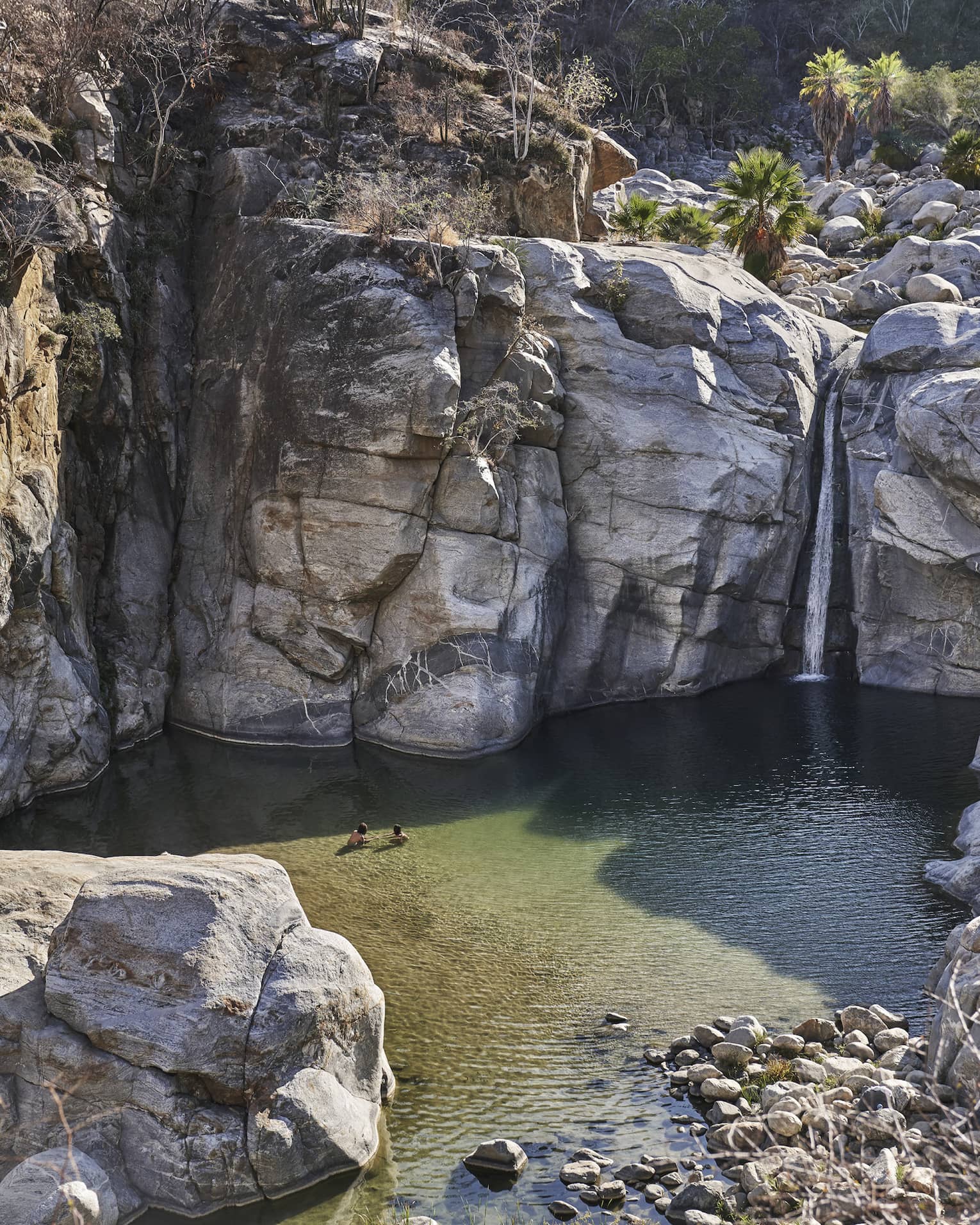 Two people wade in small ocean lagoon under large cliffs