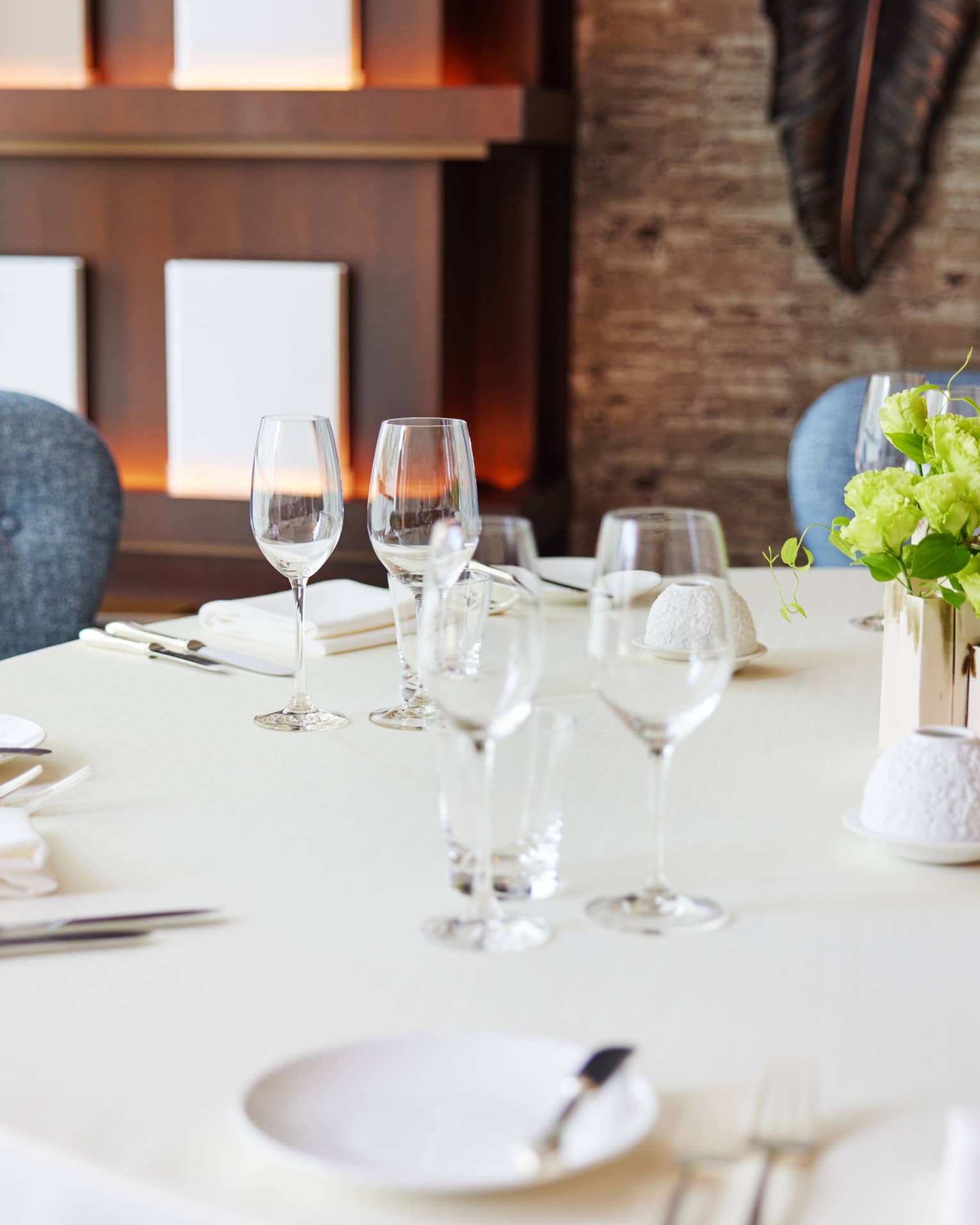 Against a warm backdrop, round table set with white tablecloth, sparkling silver and wine glasses, fresh flowers at centre.